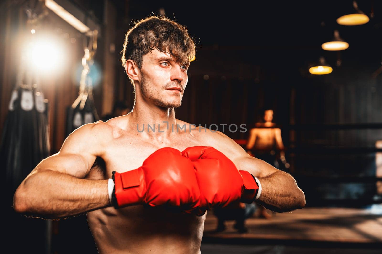 Boxing fighter posing, caucasian boxer put his hand or fist wearing glove together in front of camera in aggressive stance and ready to fight at gym with kicking bag and boxing equipment. Impetus