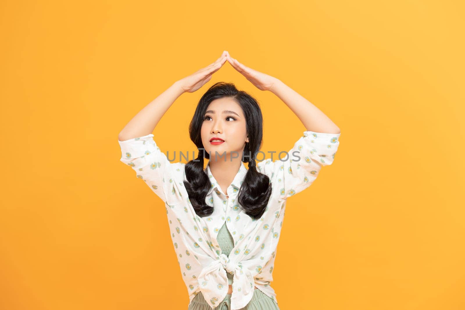 Pretty cheerful girlish girl holding hands over head like roof flat over yellow background