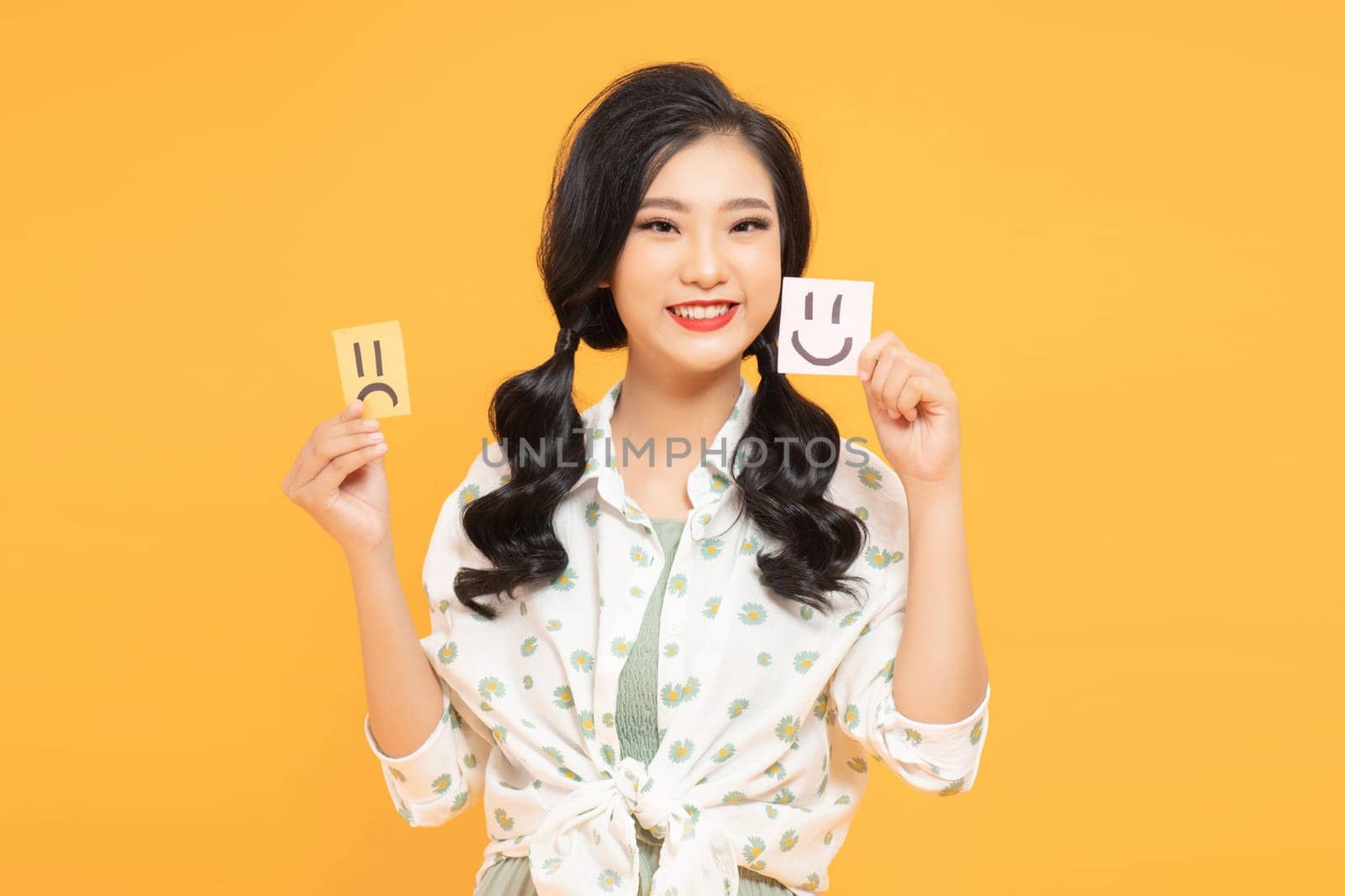 Attractive unsure girl holding in hands two draw notes isolated on yellow background by makidotvn