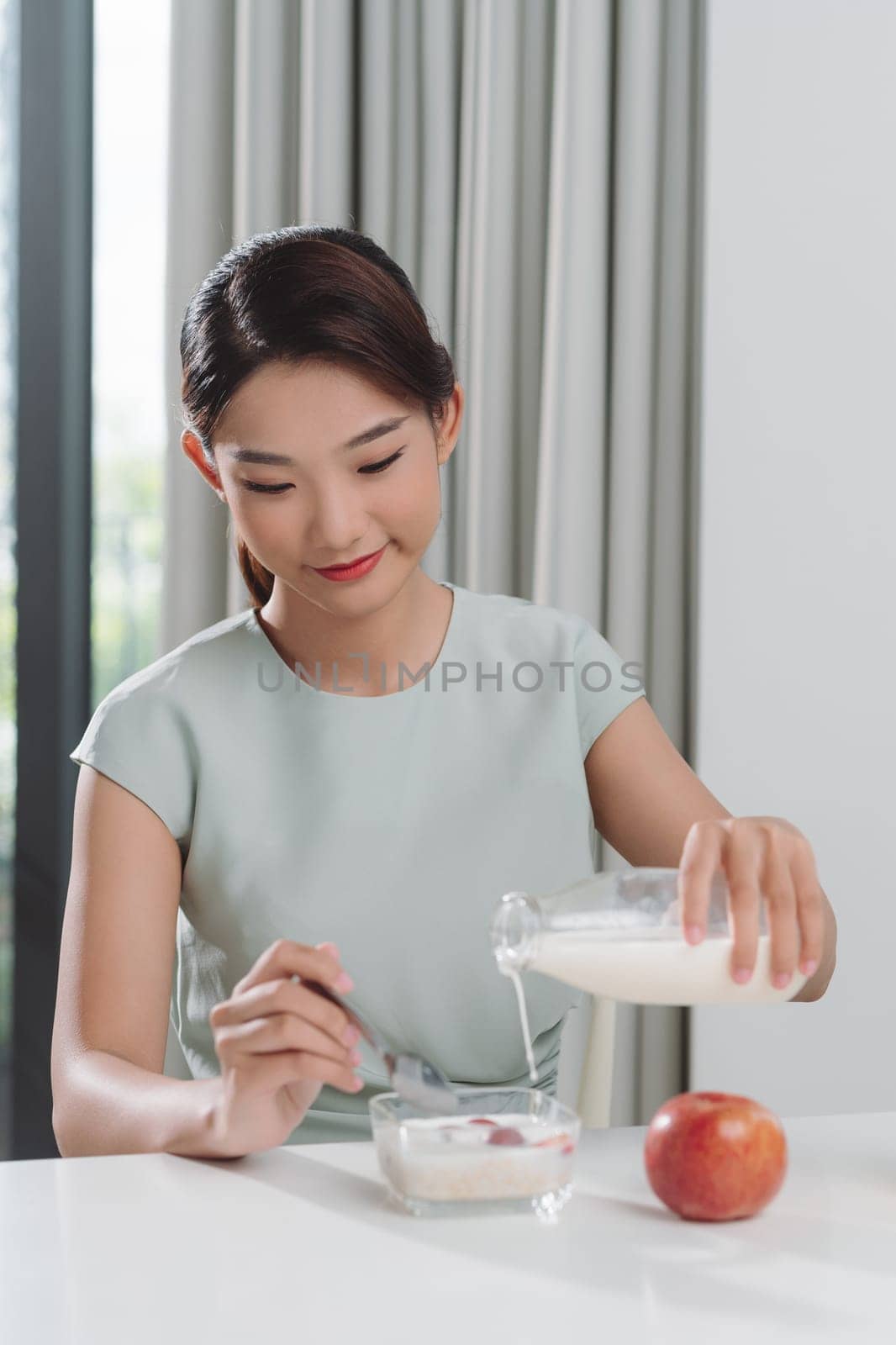 Smiling young woman pouring milk into a bowl while sitting and having breakfast  by makidotvn