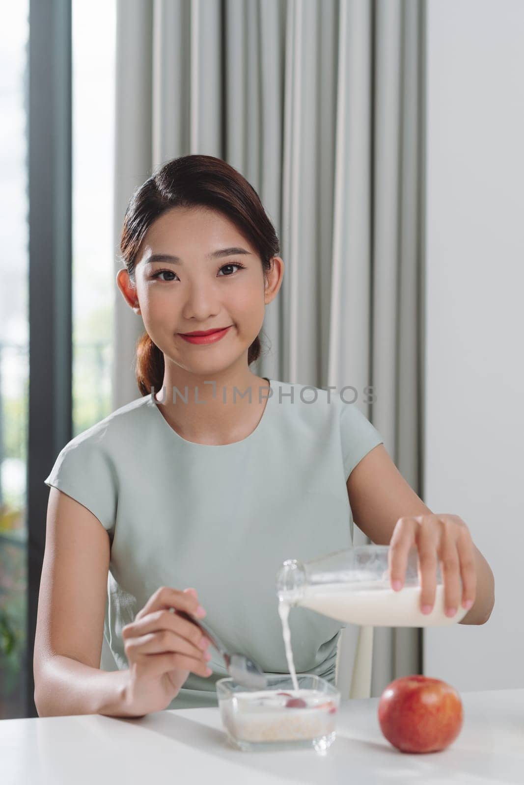 Smiling young woman pouring milk into a bowl while sitting and having breakfast 