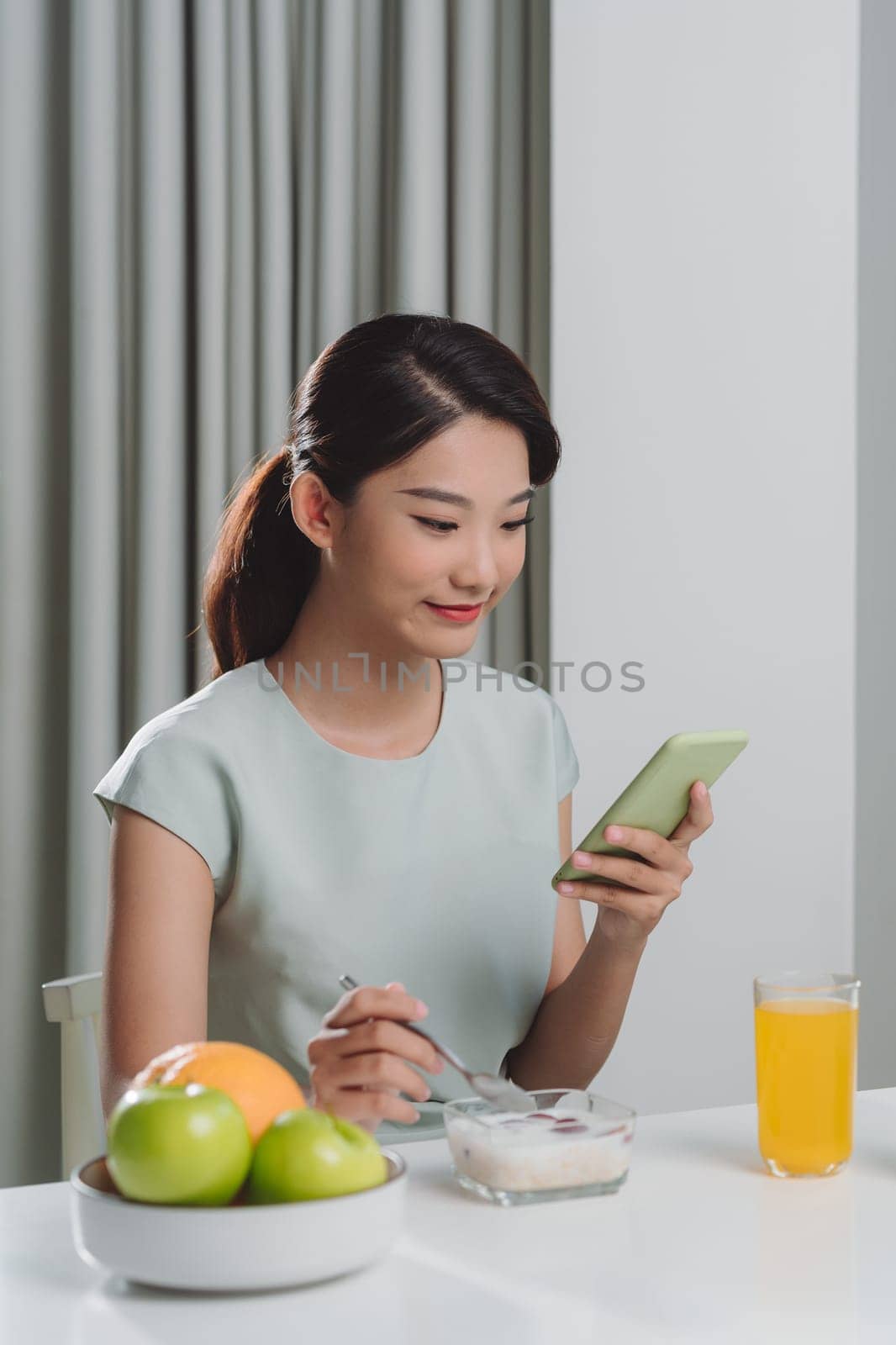 Smiling pretty asian young woman surfing on Internet while having healthy breakfast at home by makidotvn