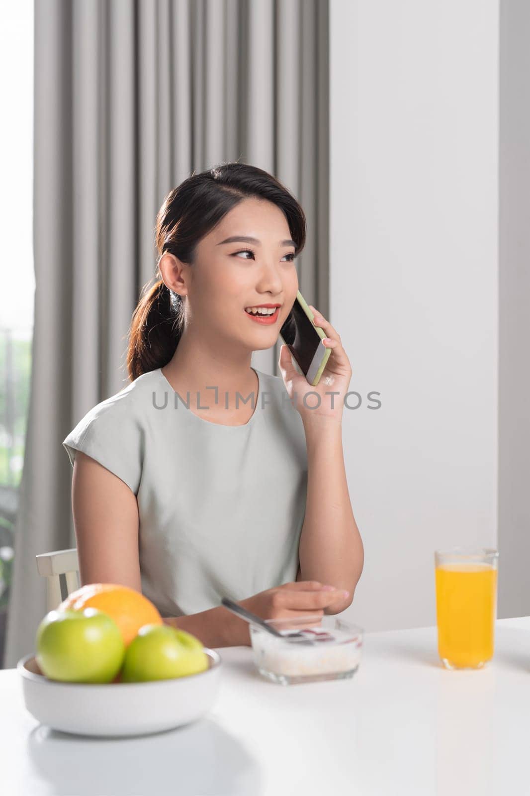 Beautiful smiling asian girl talking on phone sitting at table at dinning room in morning