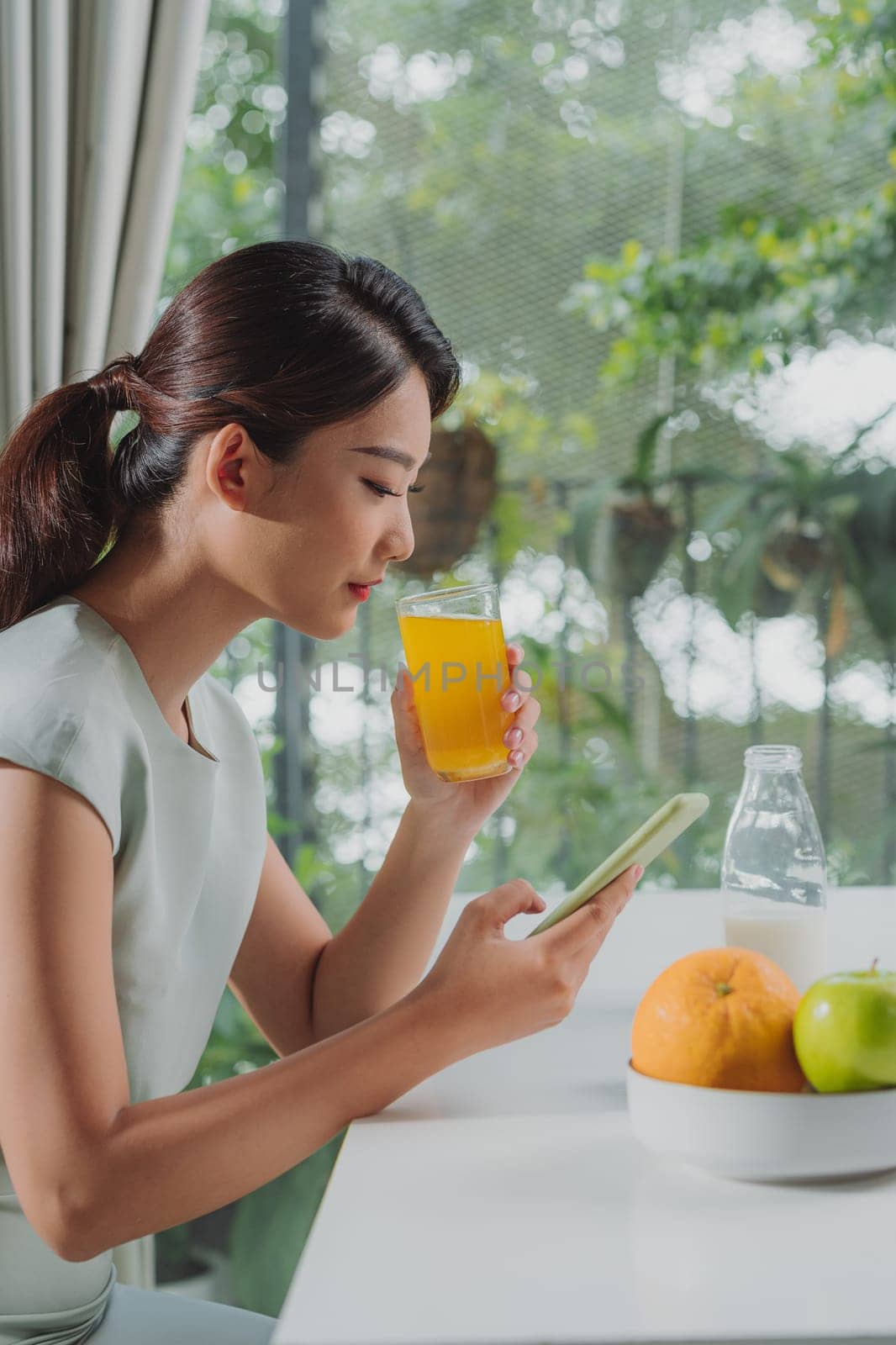 Young female using mobile phone while drinking glass of fresh orange juice during breakfast
