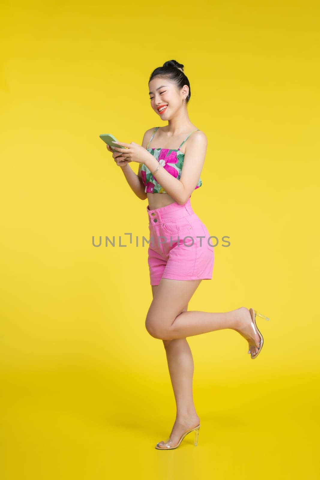 Smiling asian woman holding smartphone and looking at the camera over yellow background by makidotvn