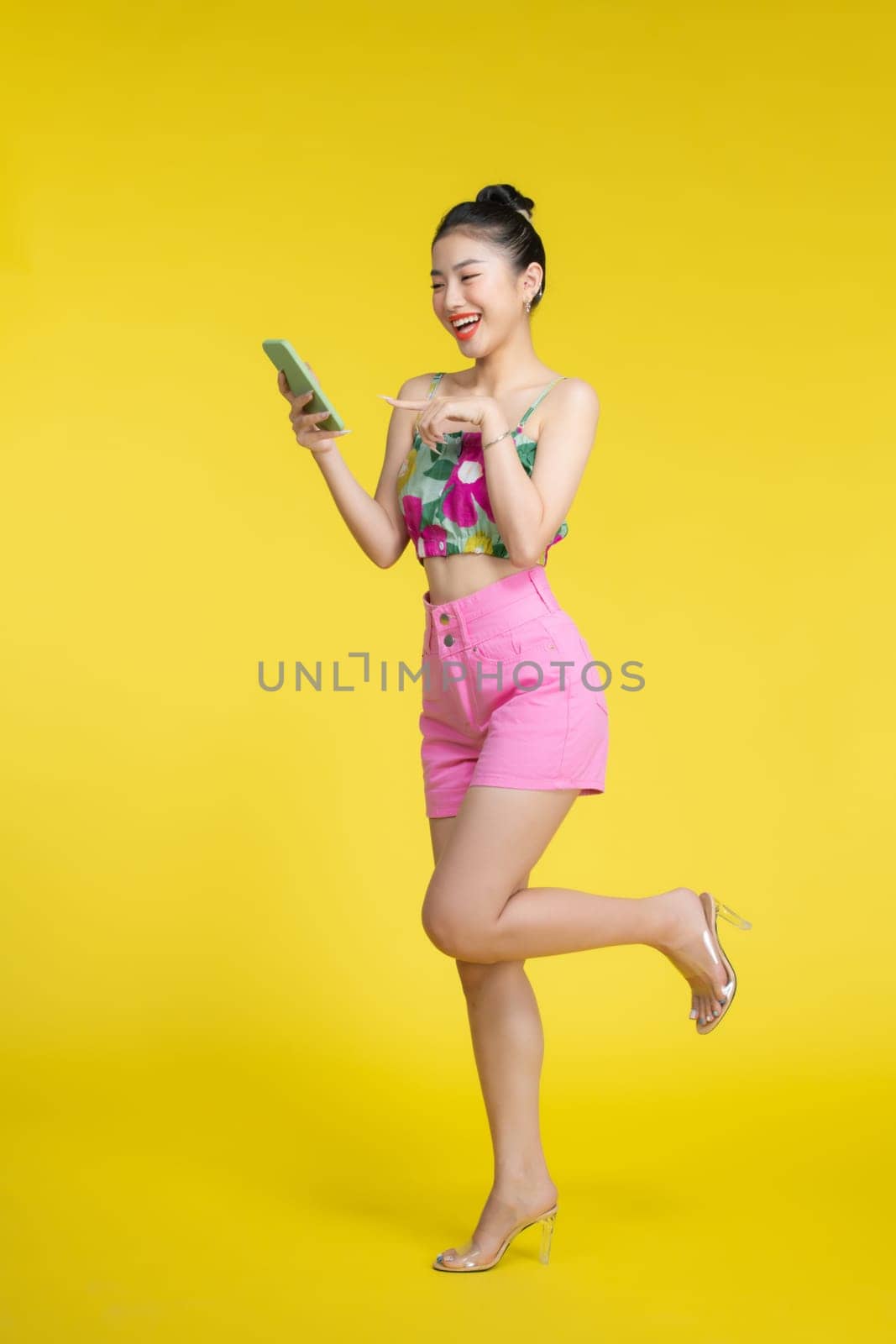 Portrait of a smiling young asian woman using mobile phone isolated over yellow background