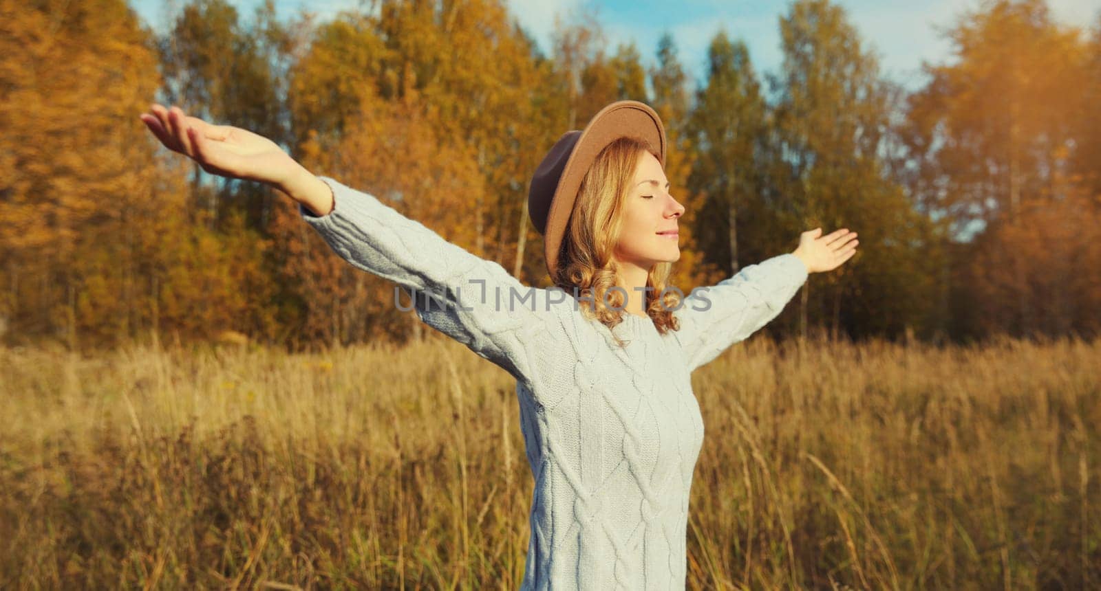 Happy smiling young woman in autumn park raising her hands up, joyful girl enjoying warm sunny weather, fall season, nature background
