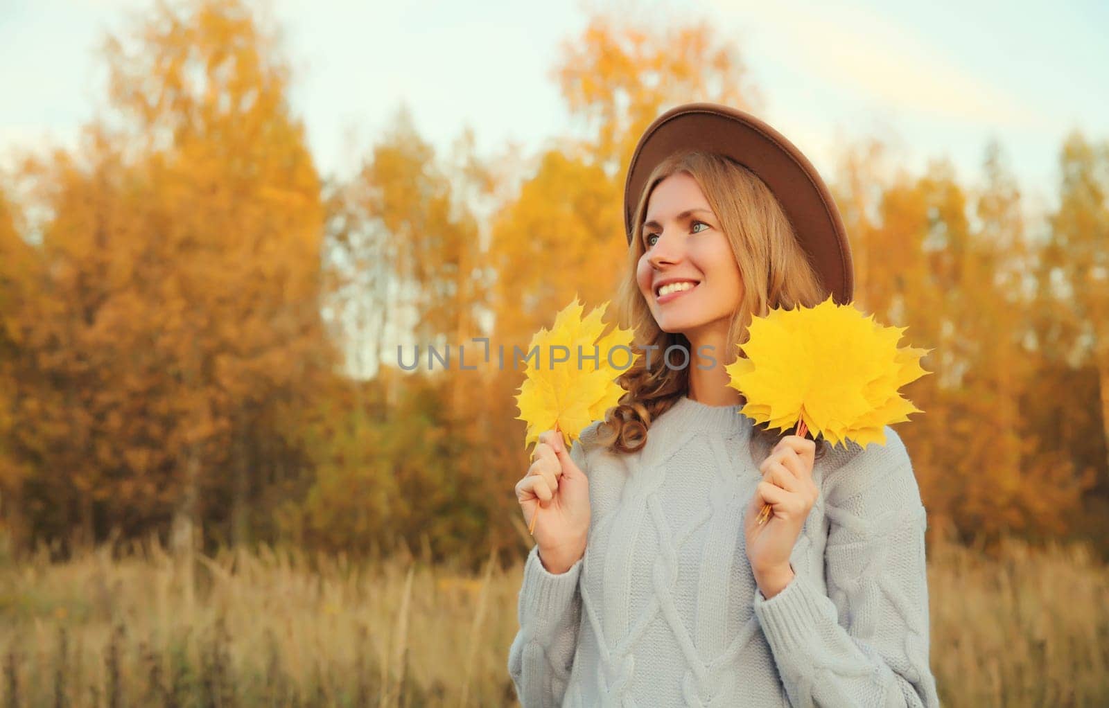 Autumn portrait of happy young woman with maple yellow leaves looking away, cheerful girl smiling wearing hat, sweater outdoors in park, warm fall season