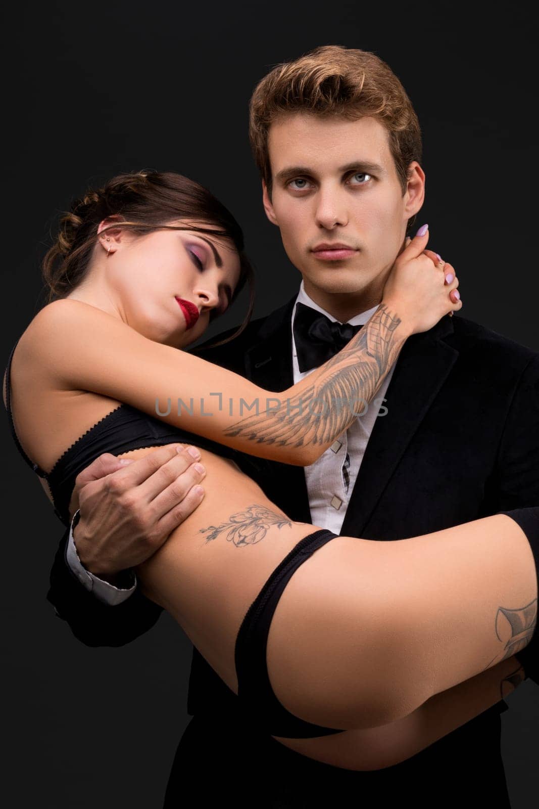 Handsome man in a suit and bowtie holds in his arms his tattooed girlfirend in black lingerie shot
