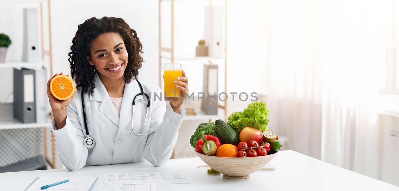 Cheerful female nutritionist african woman holding fresh orange juice and orange half in her hands while posing at modern clinic, healthy lifestyle concept