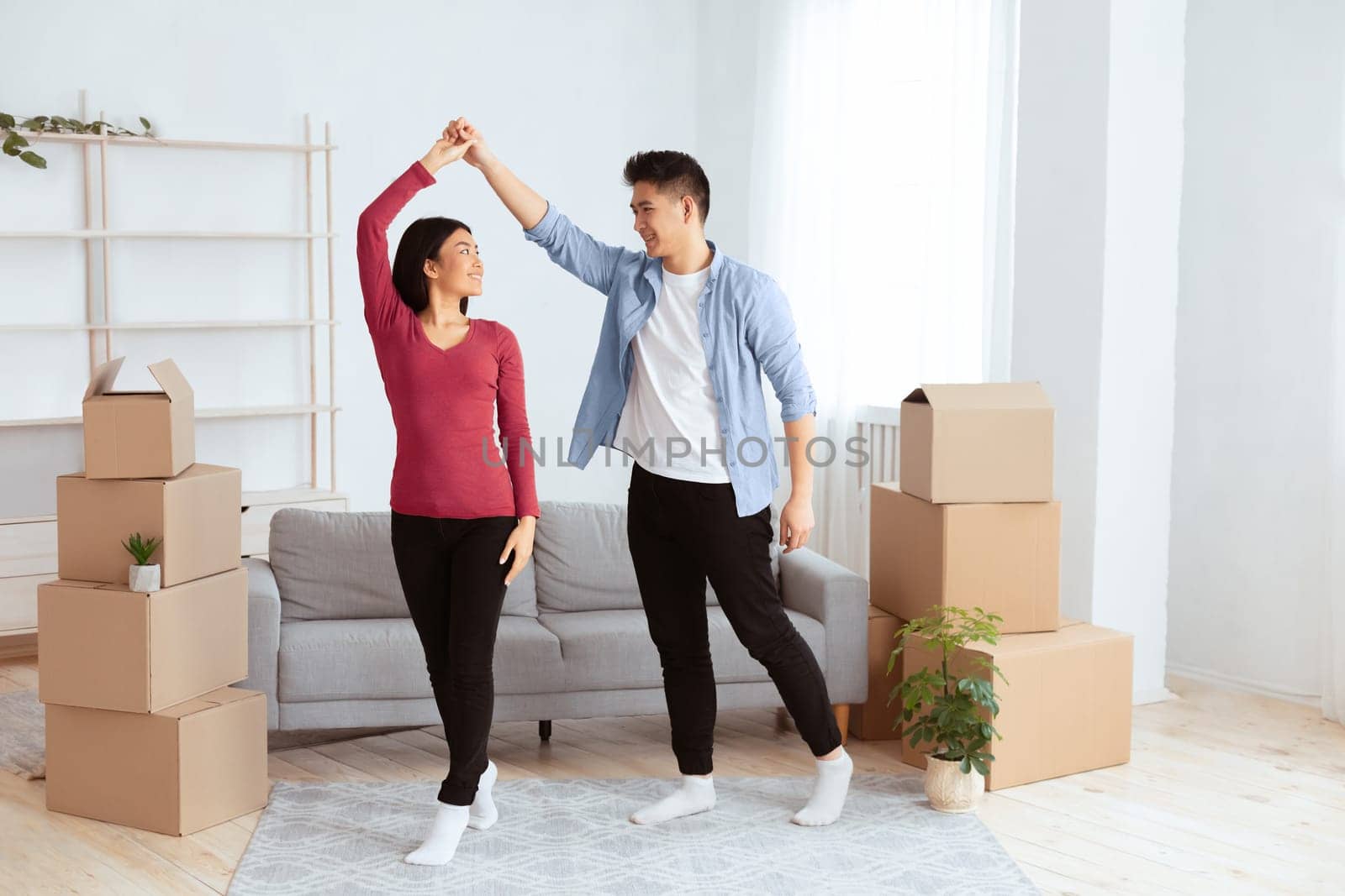 Celebrating Moving Day. Happy young asian tenants dancing among unpacked boxes in new home