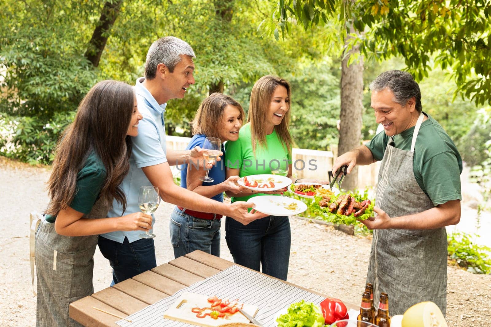 A group of five friends shares laughter and delicious food at a barbecue party in a vibrant garden. They eagerly await grilled meat while enjoying their meals and drinks.