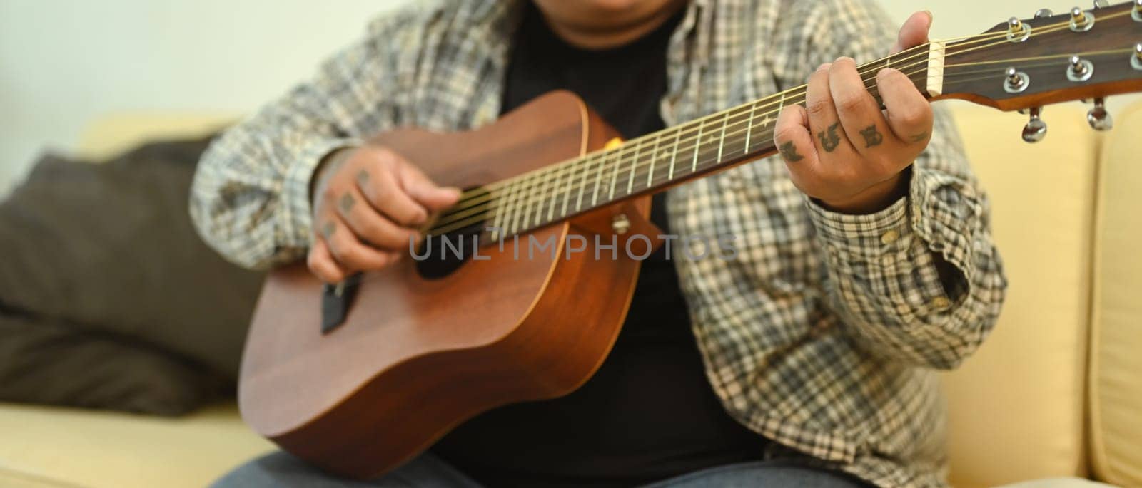 Young man playing an acoustic guitar while sitting on sofa at home. Leisure activity and hobby concept.