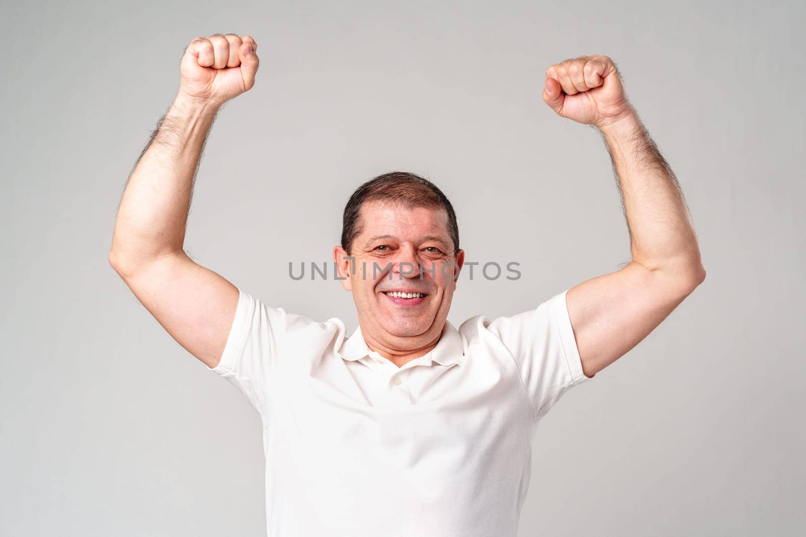 Happy older man showing off his strong biceps while standing against a light gray background close up