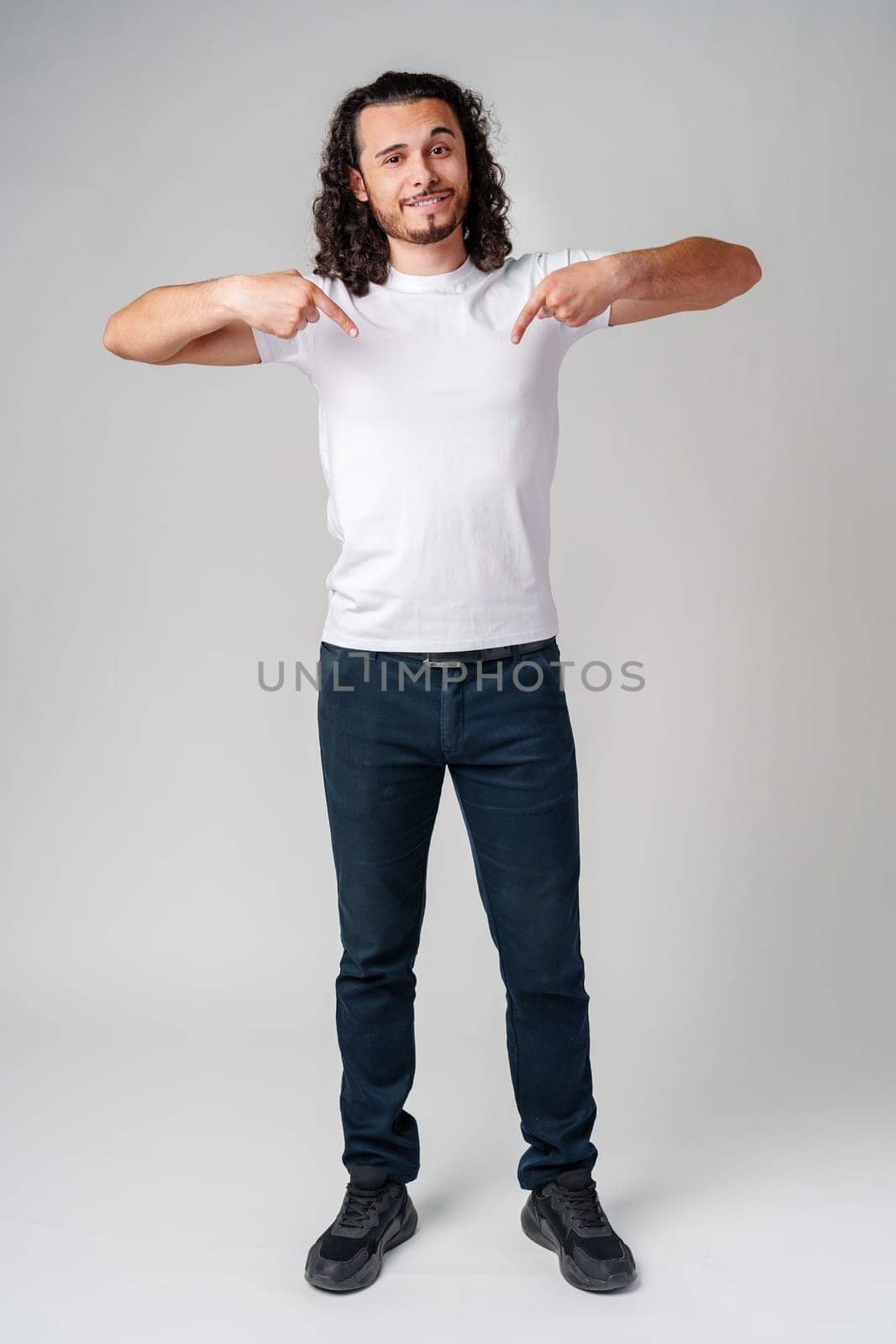A young man with curly hair joyfully pointing down while wearing a plain white t-shirt against a neutral background close up