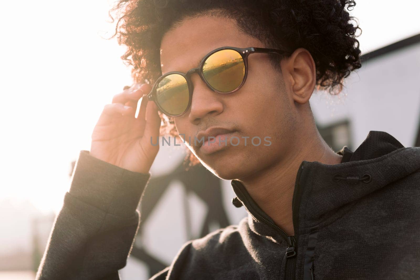 confident curly haired african american male teenager adjusting trendy sunglasses while standing against blurred urban background