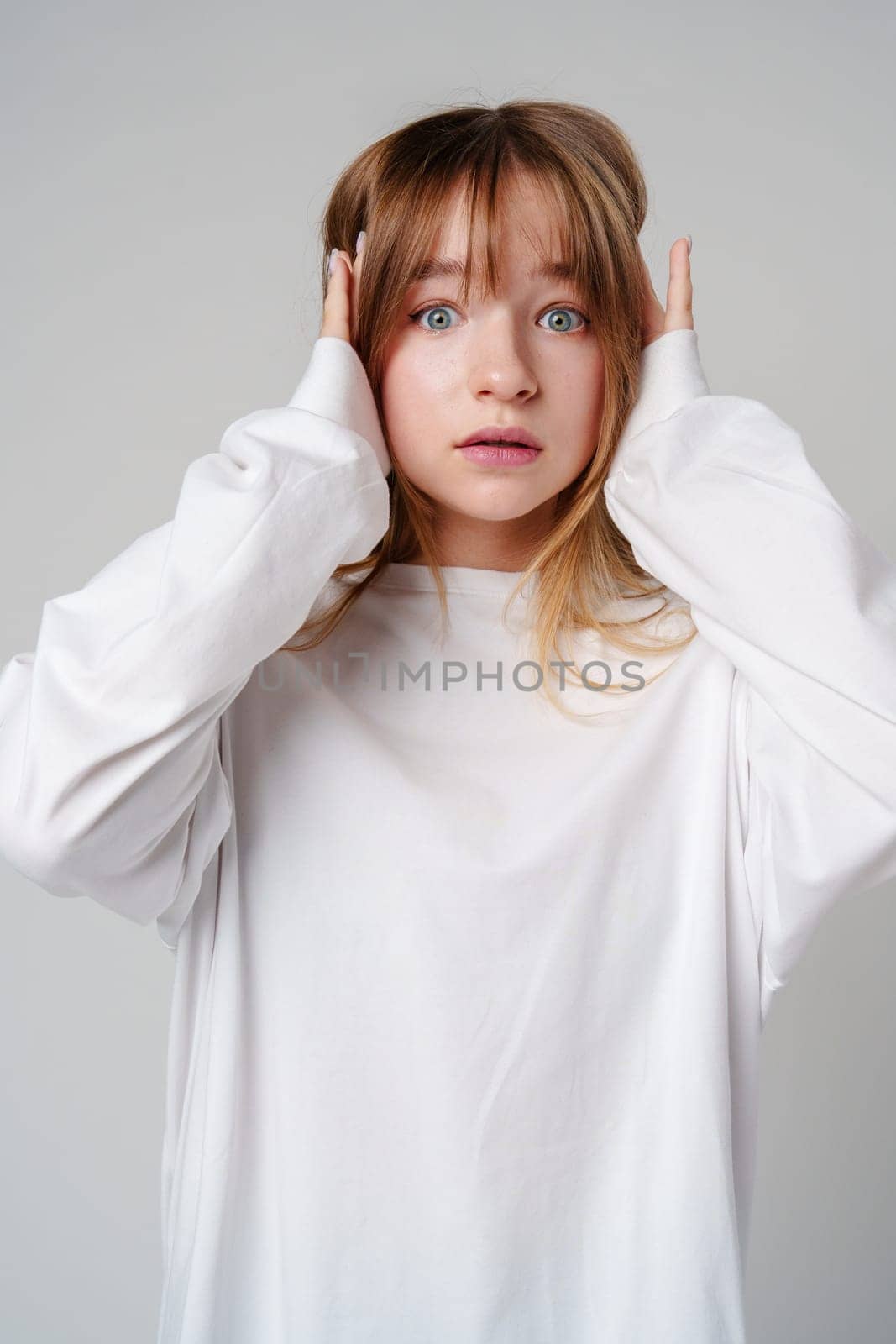 A young woman with light brown hair and blue eyes expresses surprise in a minimalist studio setting