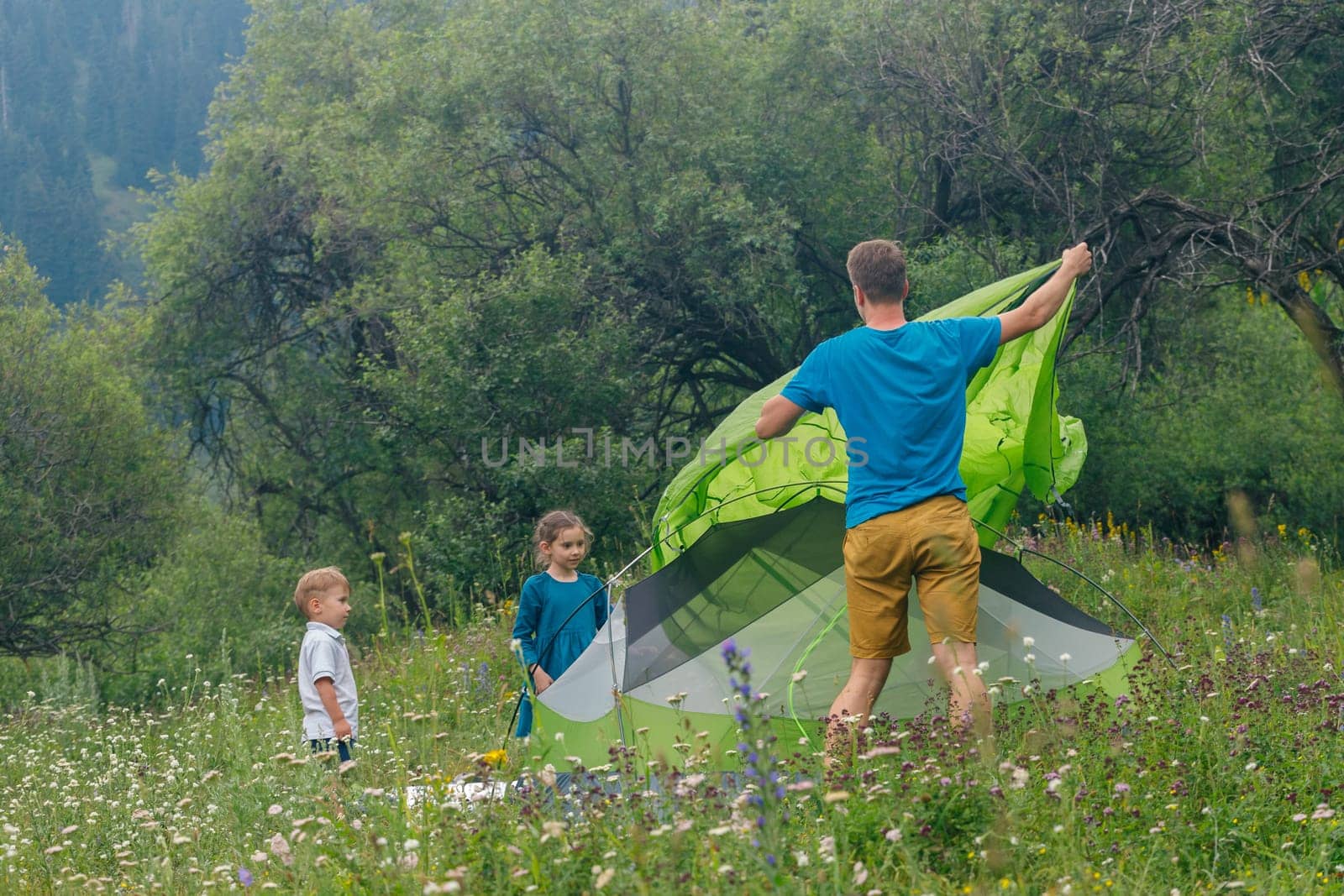 A happy father and his excited kids set up a colorful tent in a lively meadow on a sunny summer day
