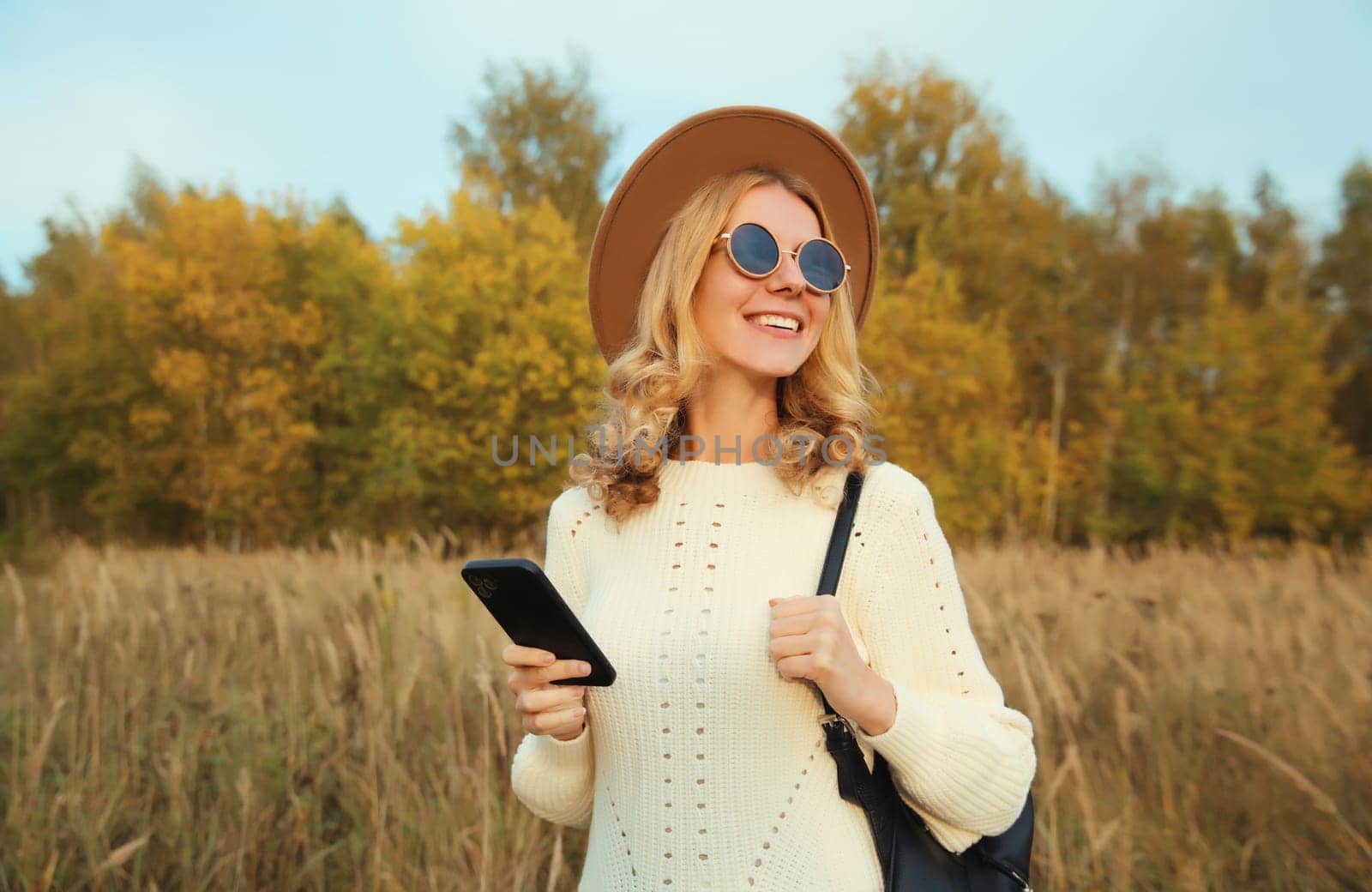 Stylish happy smiling modern young woman with phone standing in autumn park, girl holding smartphone with backpack, wear hat, sweater, glasses looking away outdoors, nature background