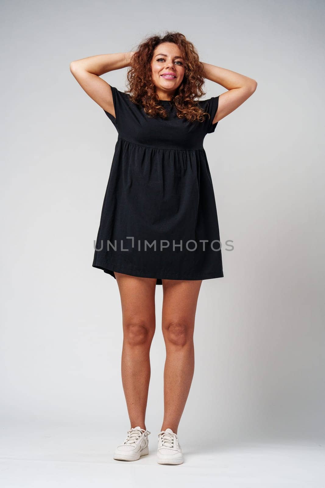 Smiling woman with curly hair poses confidently against a plain background while holding her hands behind her head indoors