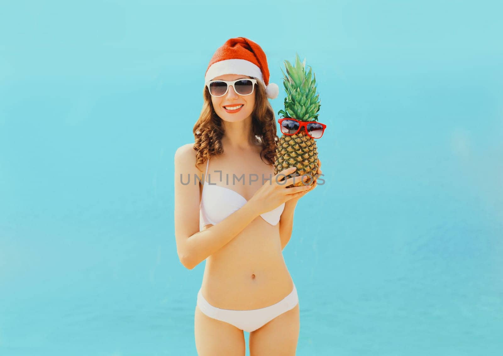 Christmas portrait happy woman in red santa hat with pineapple on beach over sea background