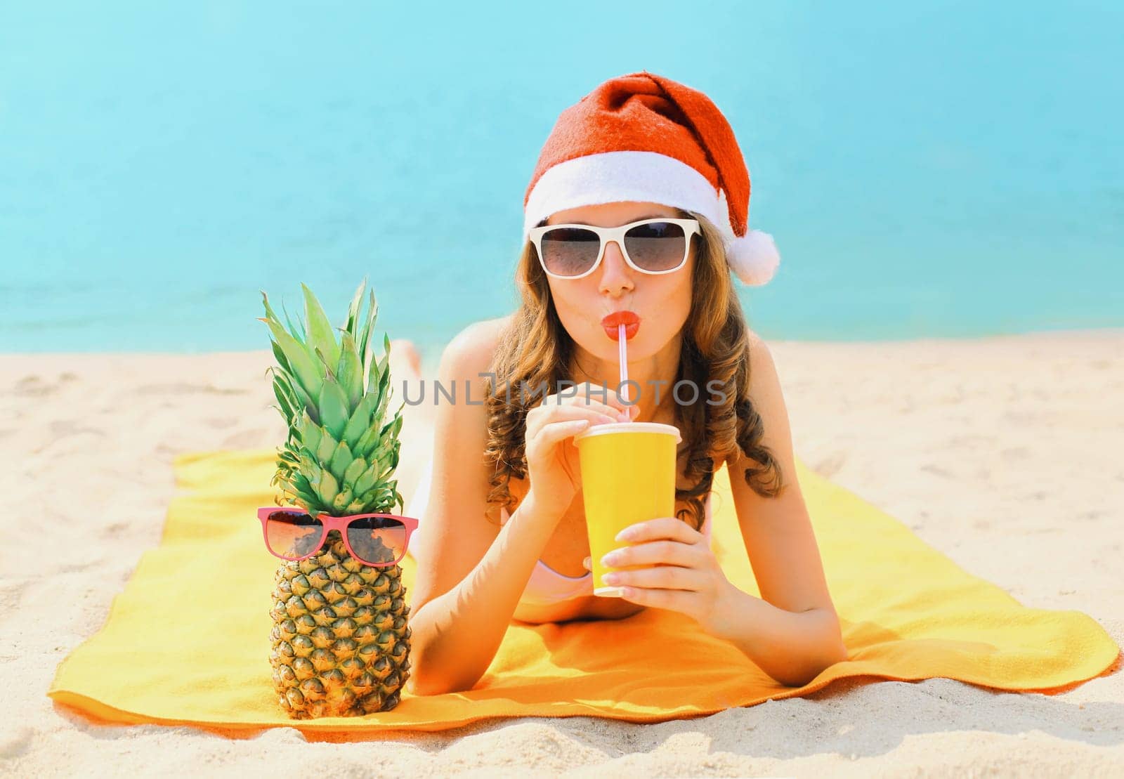 Christmas portrait woman in red santa hat with pineapple drinking juice lying on beach on sea