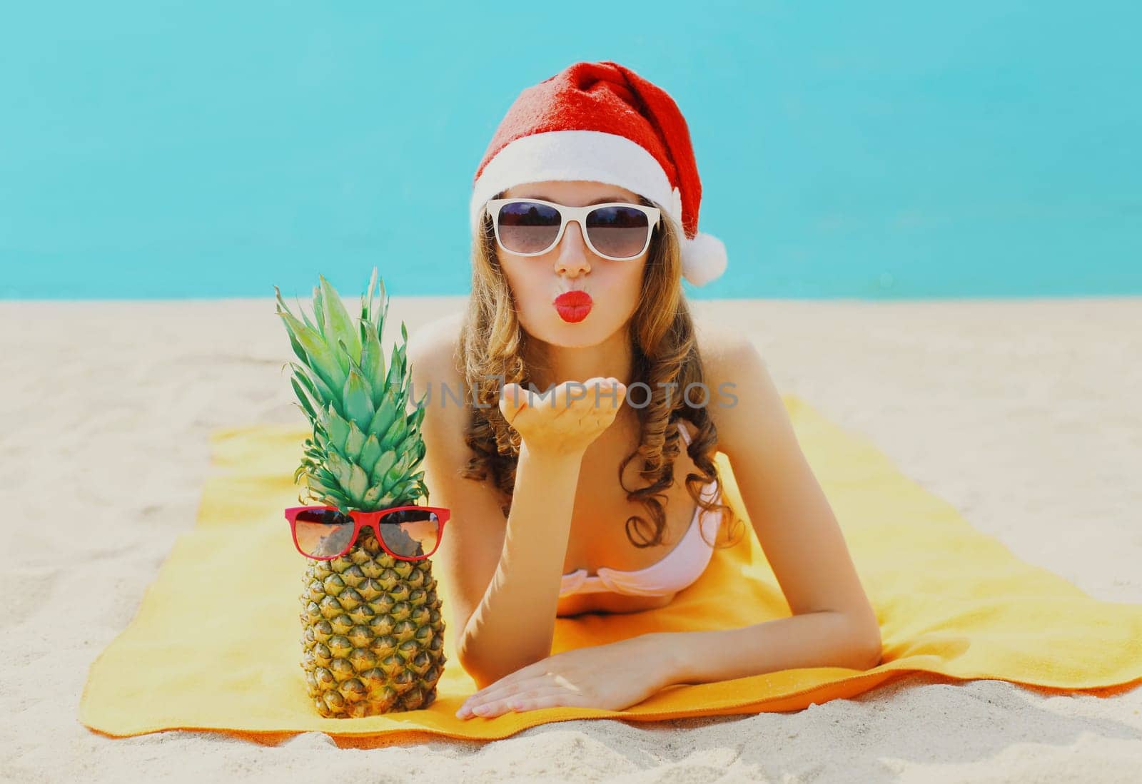 Christmas portrait woman in red santa hat with pineapple lying on beach blowing kiss on sea