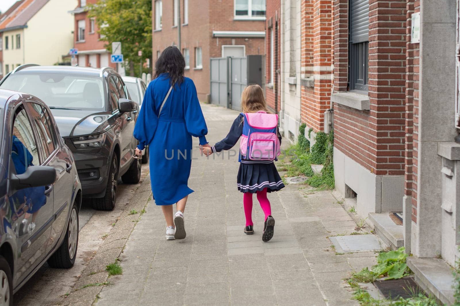 Mother and teenage daughter walking home from school to parking lot to car to go home and do homework, girl in school blue uniform with pink backpack, view from back, education concept, back to school