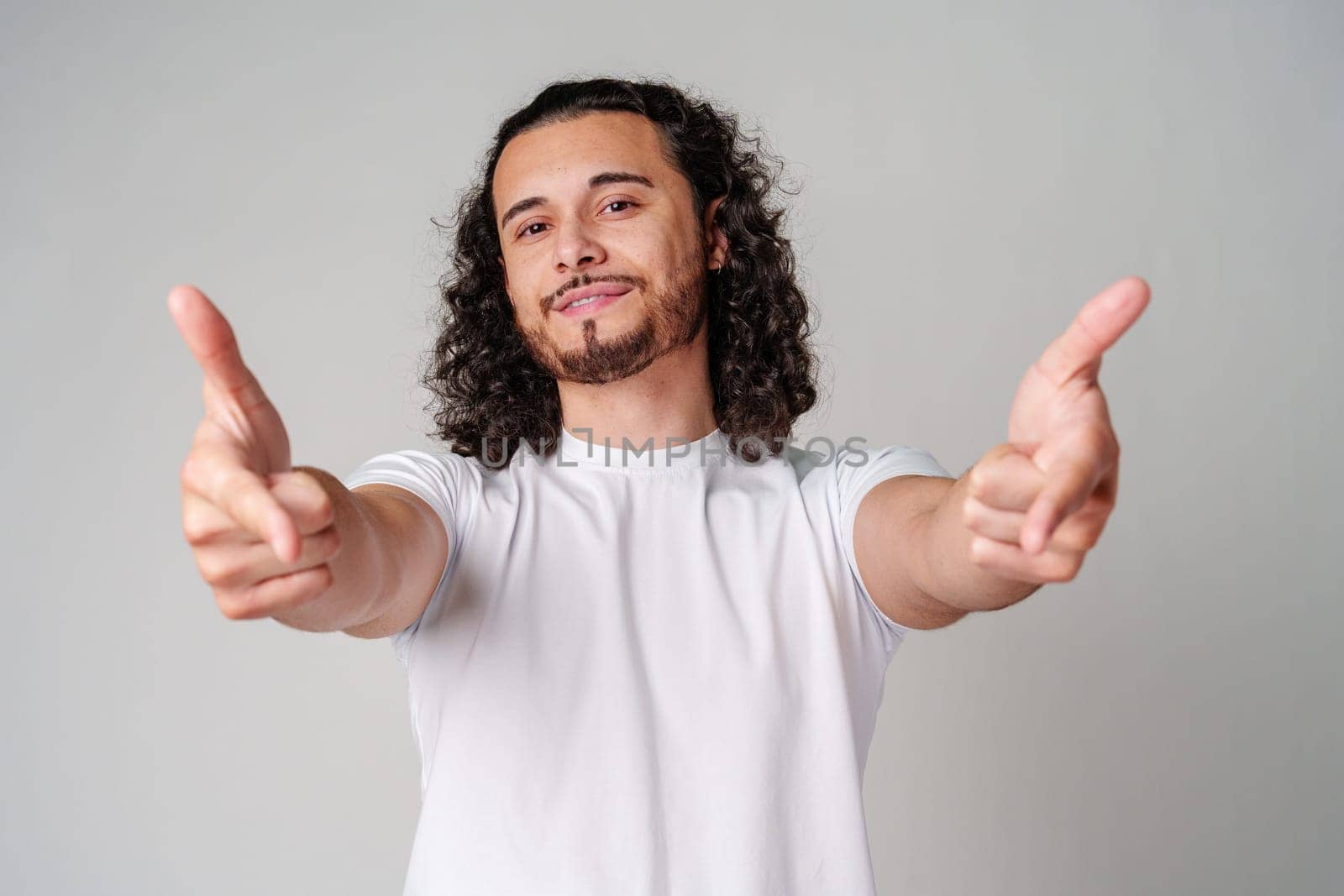 A smiling young man with curly hair playfully pointing directly at the viewer in a light-colored setting close up