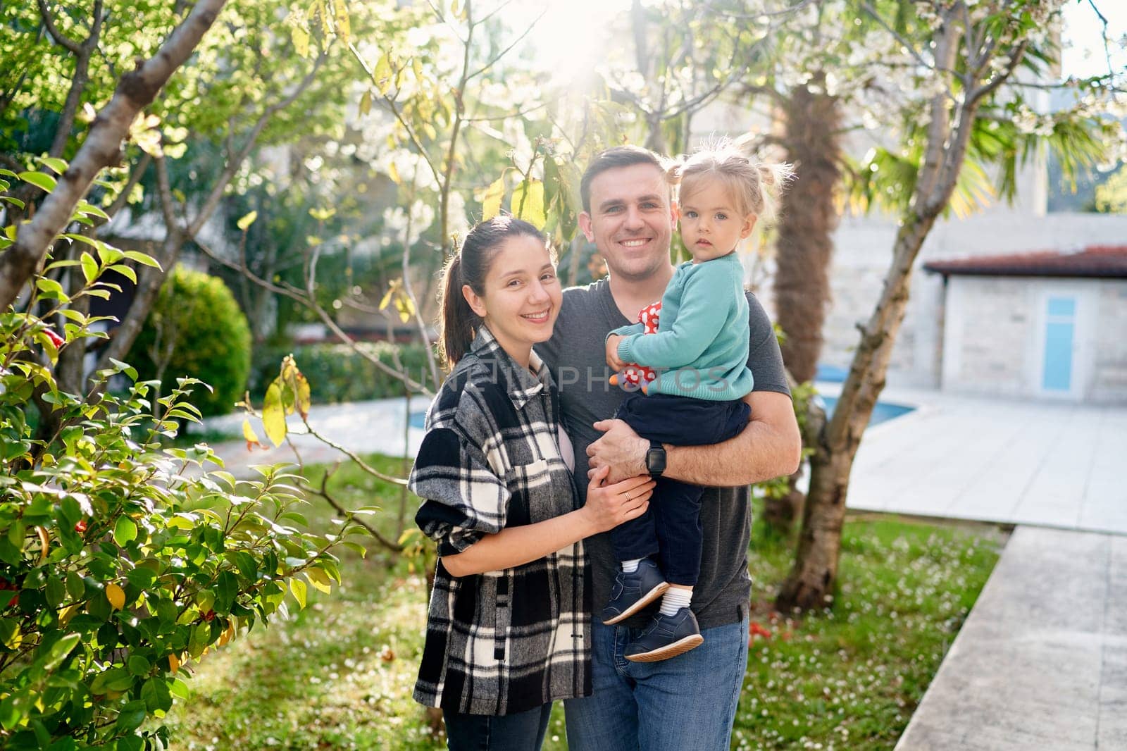 Smiling mother hugs father with a little girl in his arms while standing in a blooming garden near the house. High quality photo