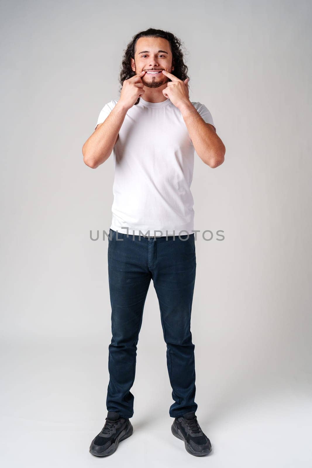 Young man in a white t-shirt playfully smiles while pointing to his teeth against a plain gray background in a studio setting