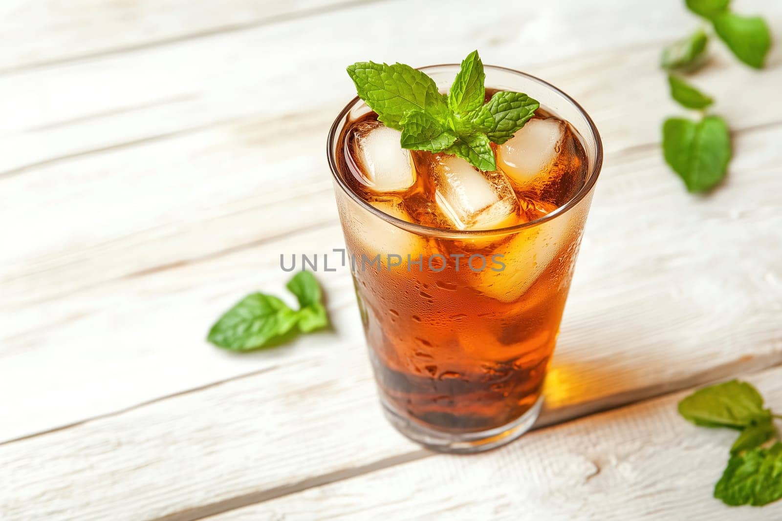 A tall glass filled with cold iced beverage and ice cubes is garnished with fresh mint leaves, placed on a rustic wooden table in the afternoon light.