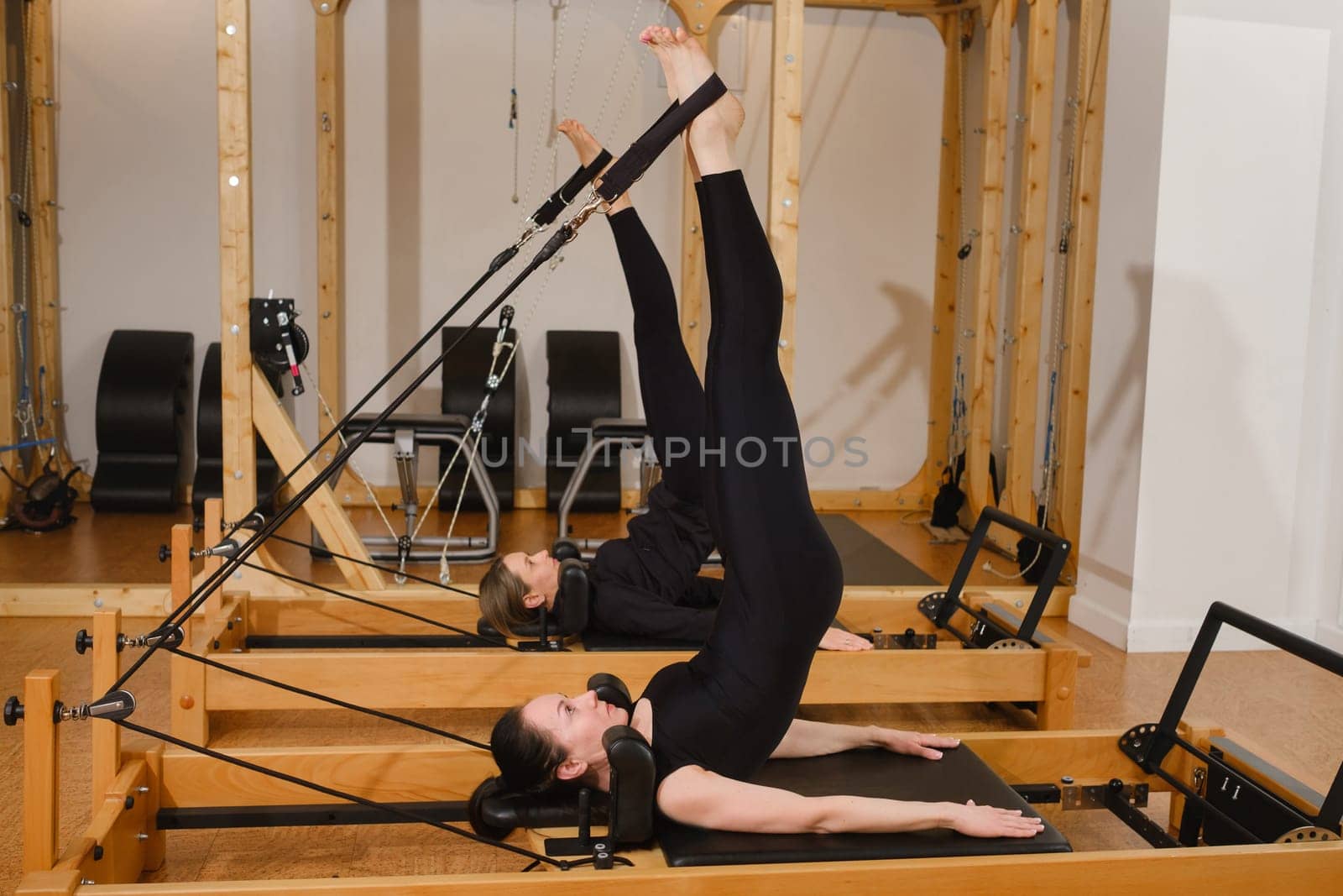Two women in black sportswear are doing pilates on a reformer machine in the gym.