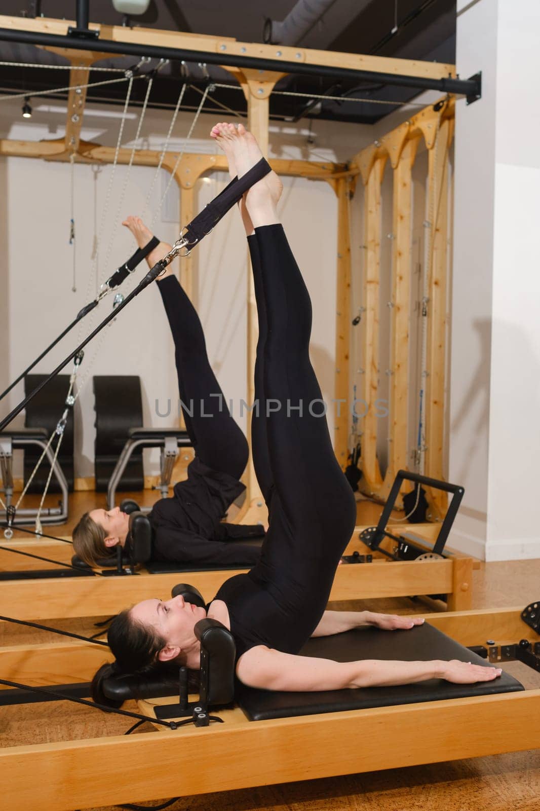 Two women in black sportswear are doing pilates on a reformer machine in the gym.