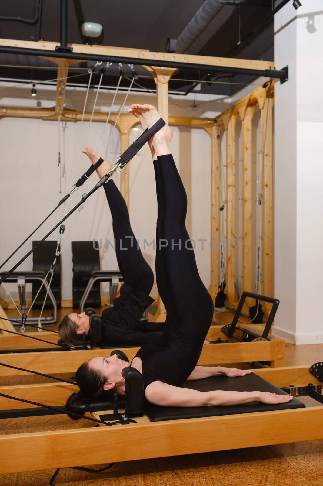 Two women in black sportswear are doing pilates on a reformer machine in the gym.