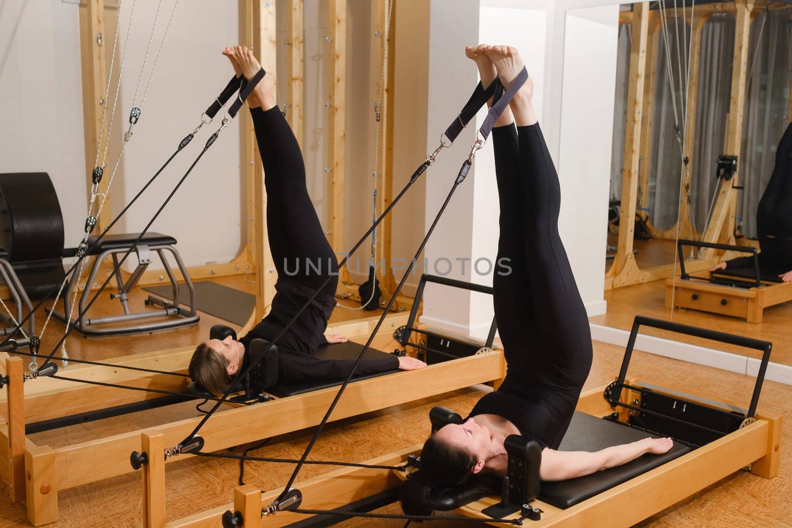 Two women in black sportswear are doing pilates on a reformer machine in the gym.