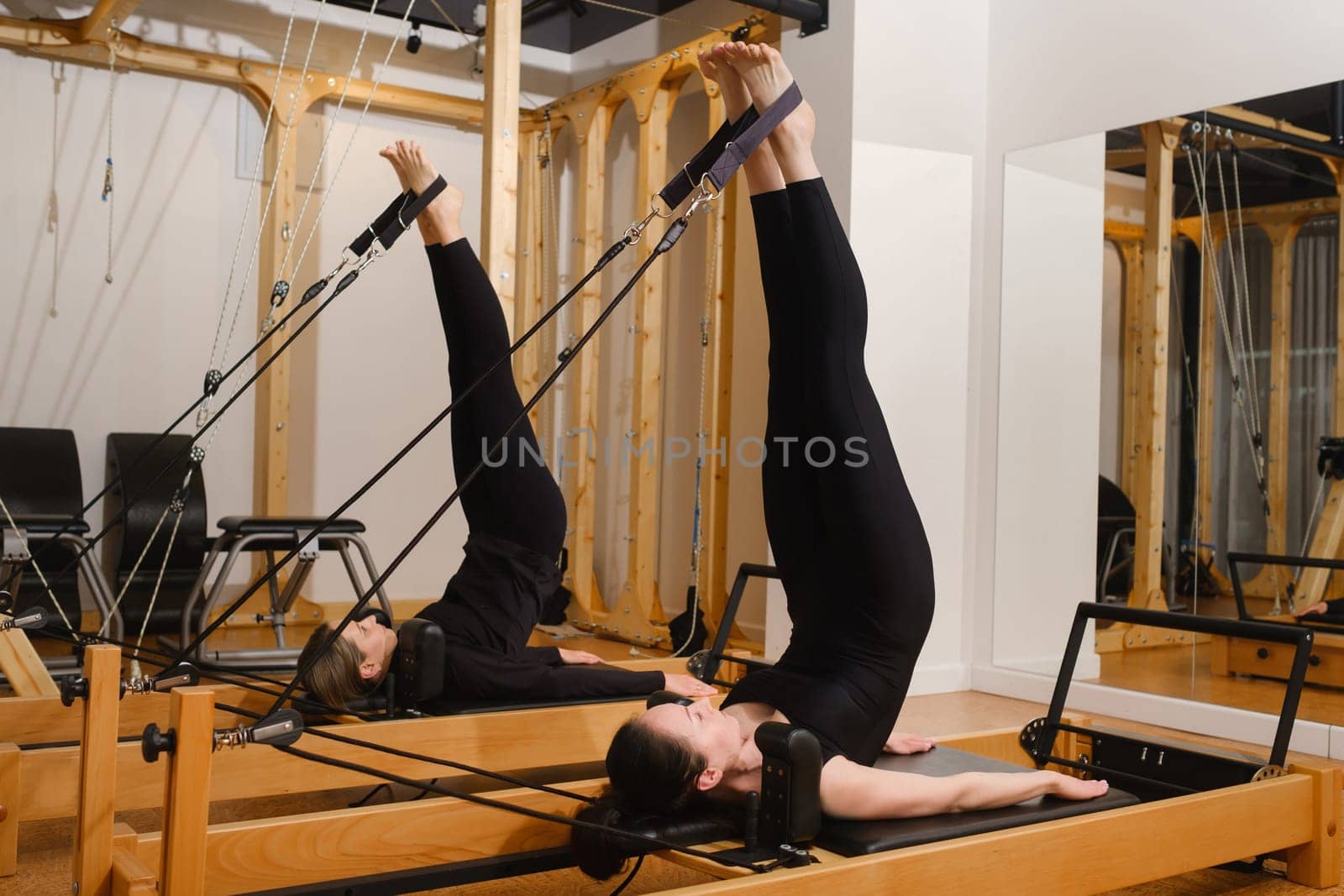 Two women in black sportswear are doing pilates on a reformer machine in the gym.