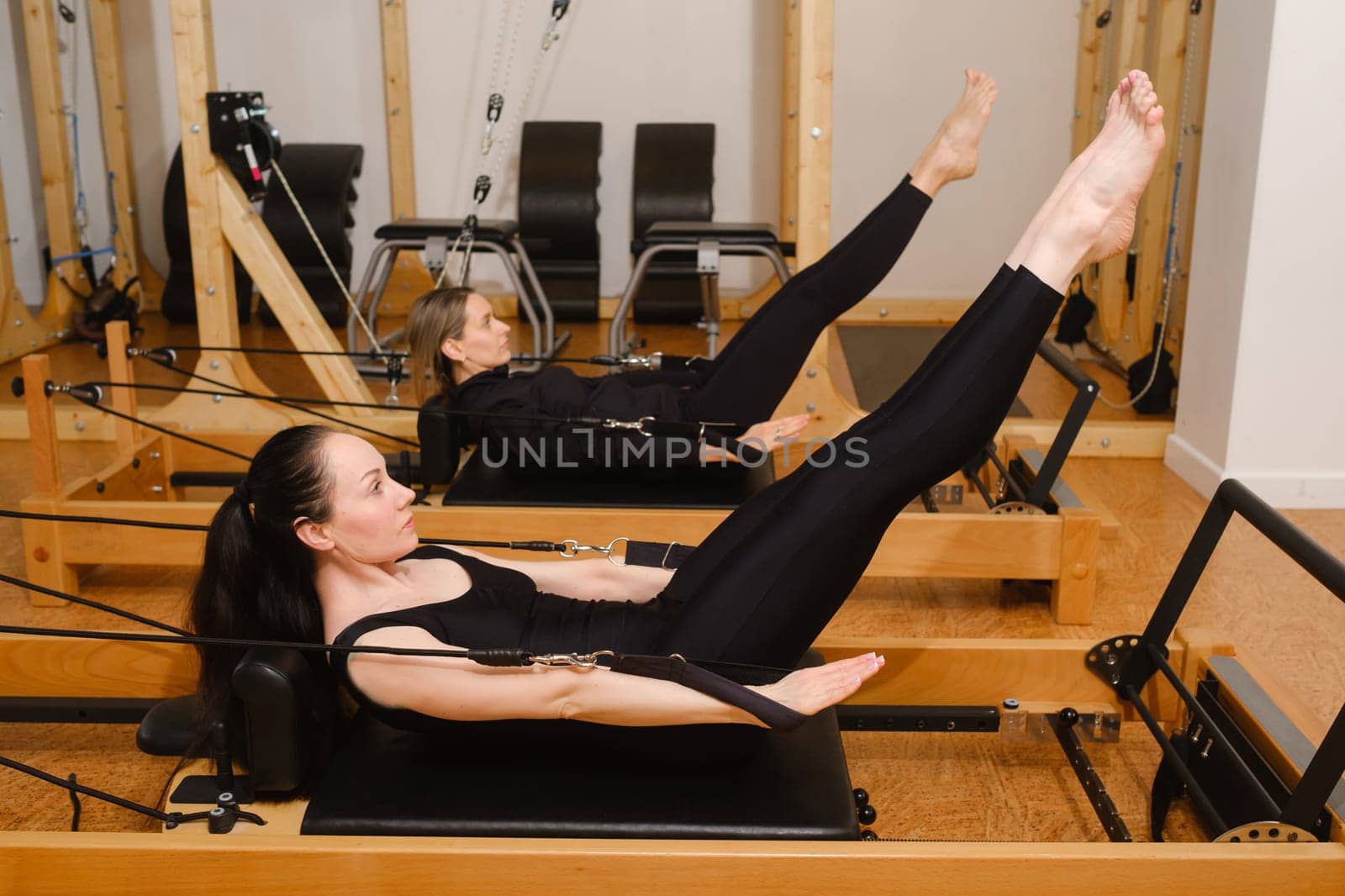 Two women in black sportswear are doing pilates on a reformer machine in the gym.
