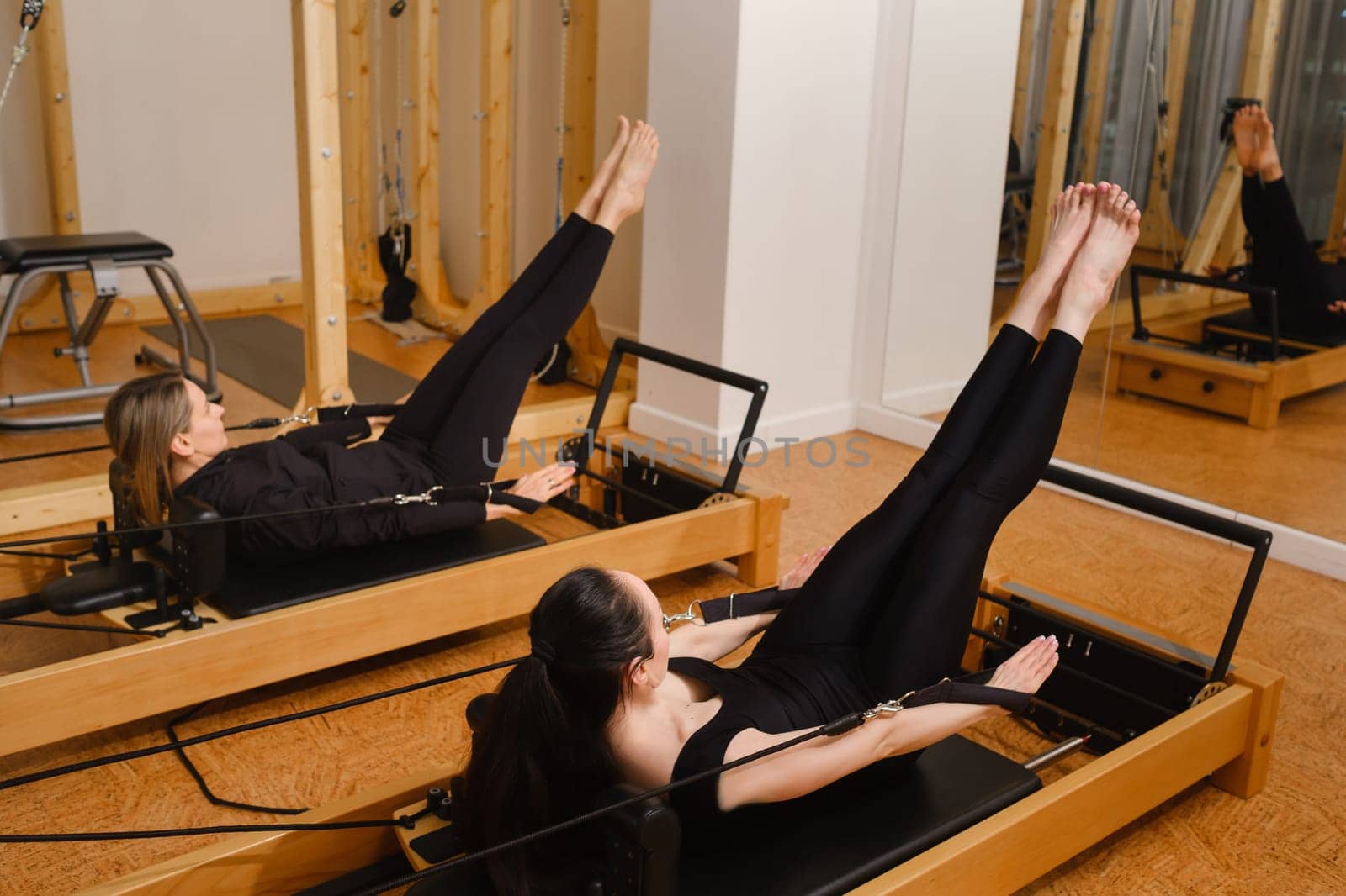 Two women in black sportswear are doing pilates on a reformer machine in the gym.