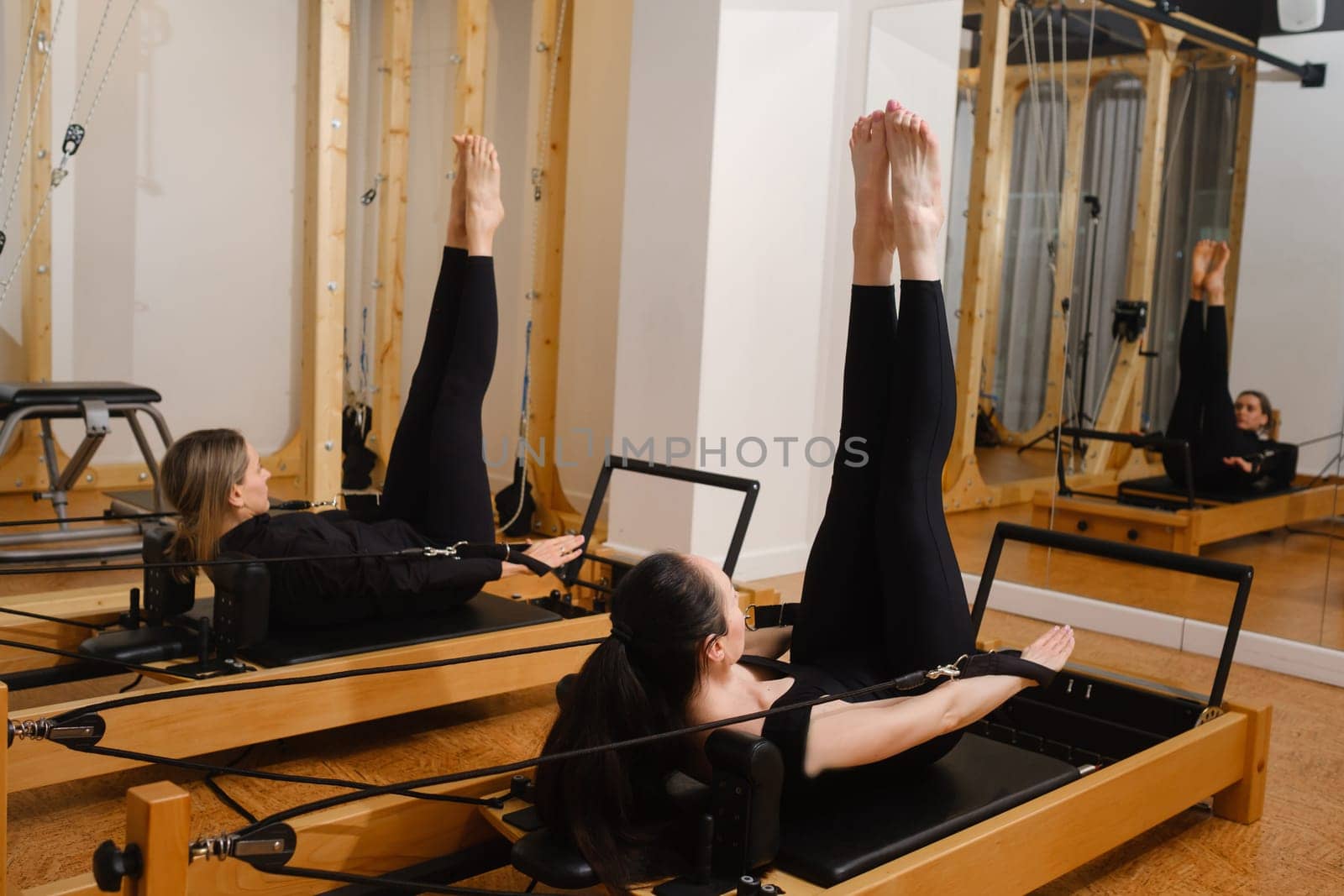 Two women in black sportswear are doing pilates on a reformer machine in the gym.