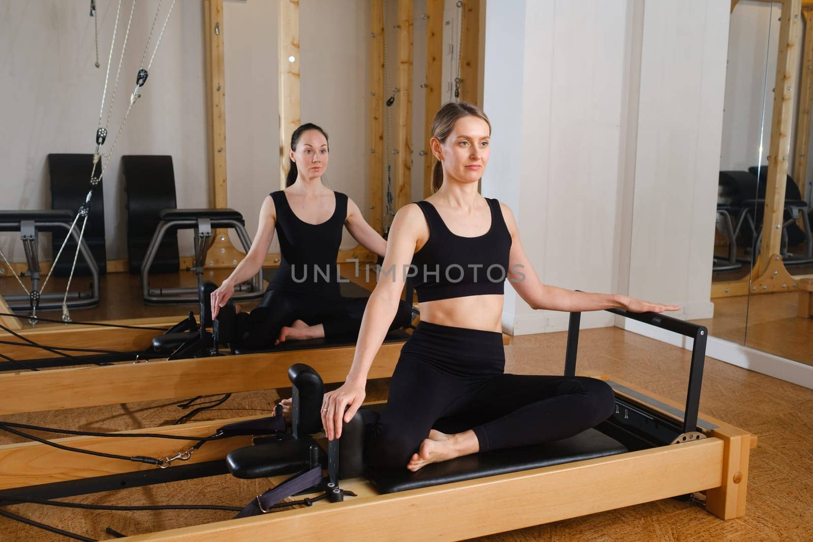 Two women sitting on a reformer exercise machine perform an exercise in the gym.