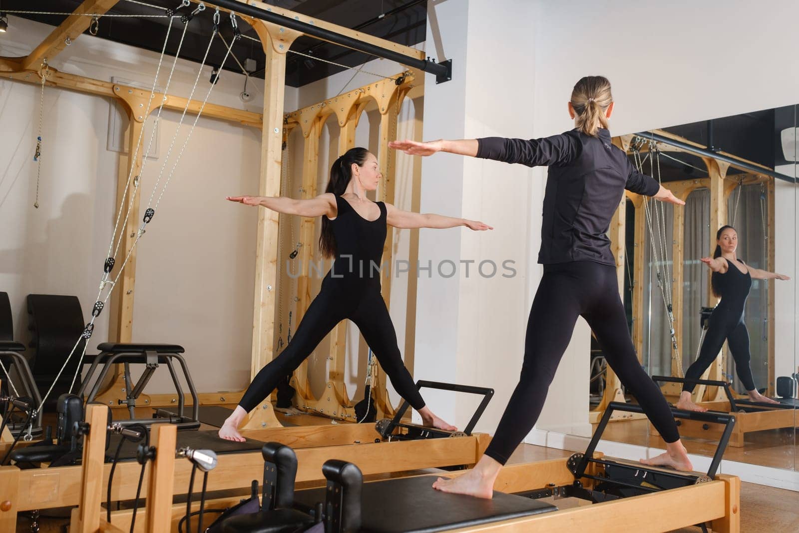 Two women standing on a reformer exercise machine perform an exercise in the gym.