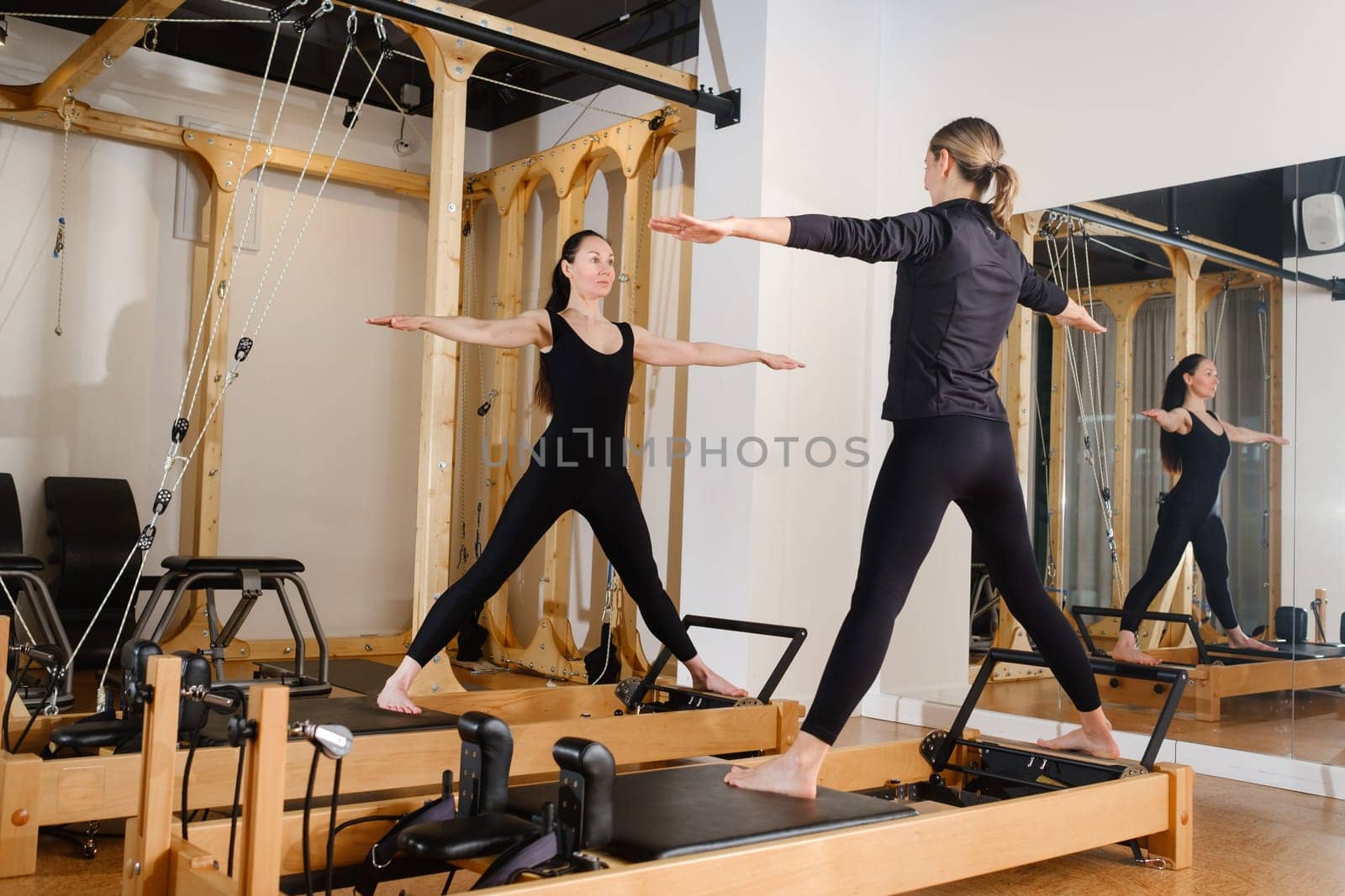 Two women standing on a reformer exercise machine perform an exercise in the gym.