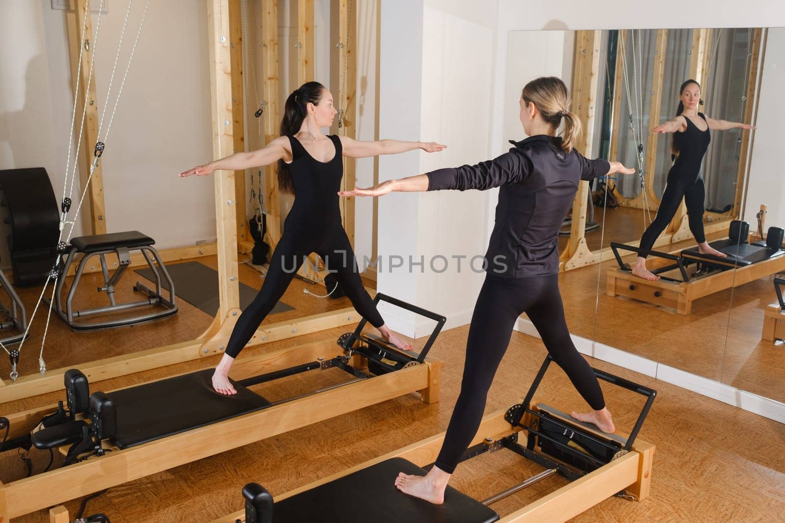 Two women standing on a reformer exercise machine perform an exercise in the gym.