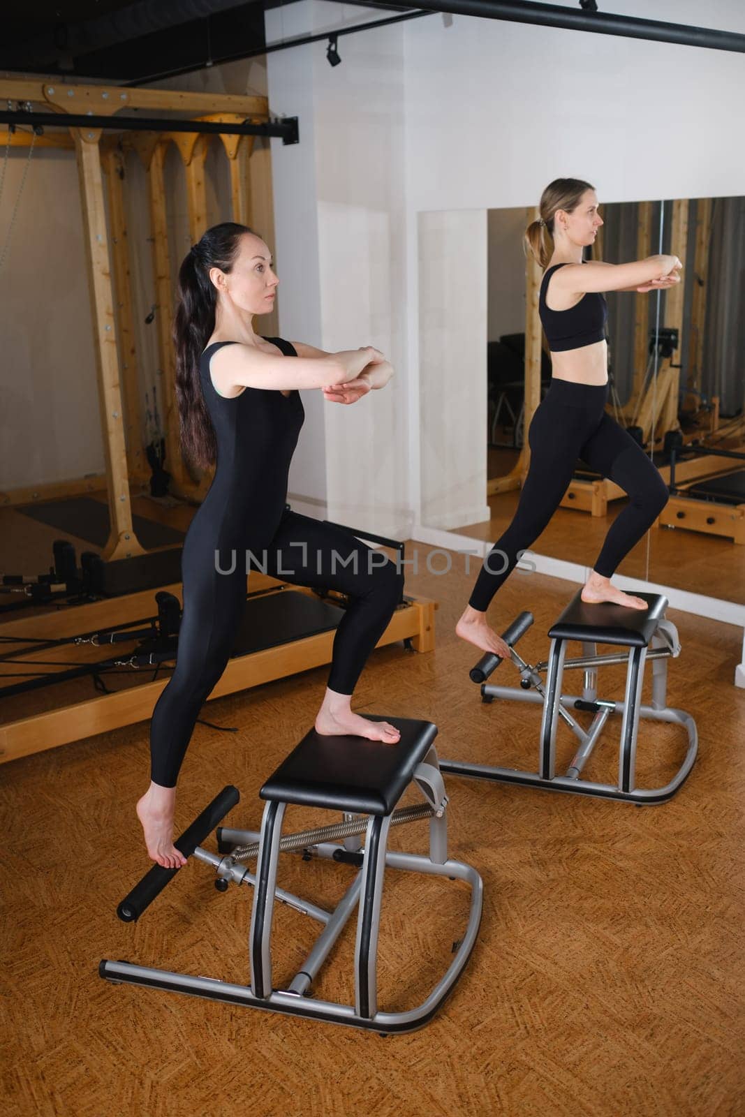 Two girls doing Pilates on a Pilates chair in a Pilates gym. A beautiful slim woman in black clothes performs exercises to strengthen the spine and leg muscles.