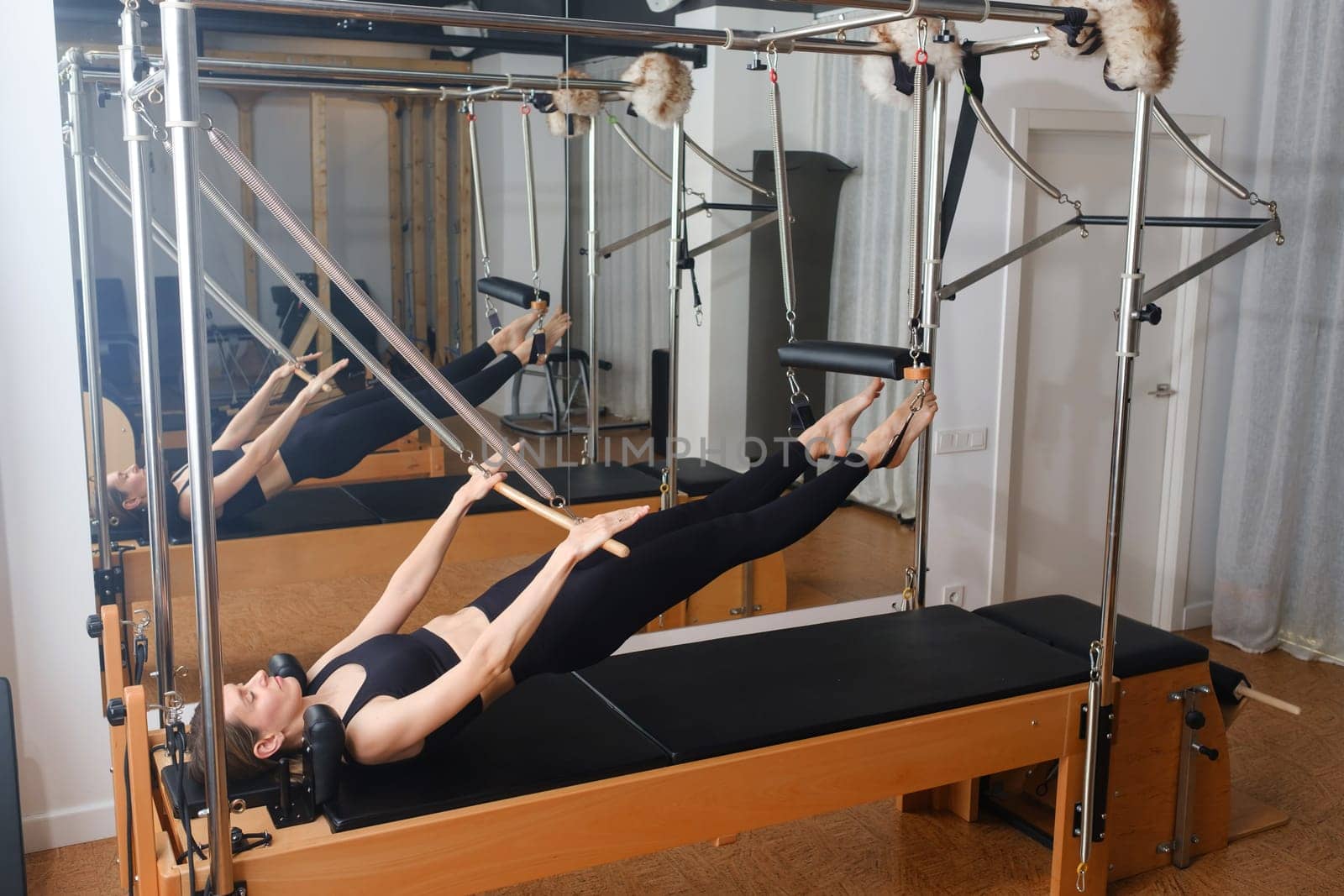 Young girl doing Pilates on trapeze table in the gym. Beautiful slim woman in black clothes performs exercises to strengthen the spine and back muscles.