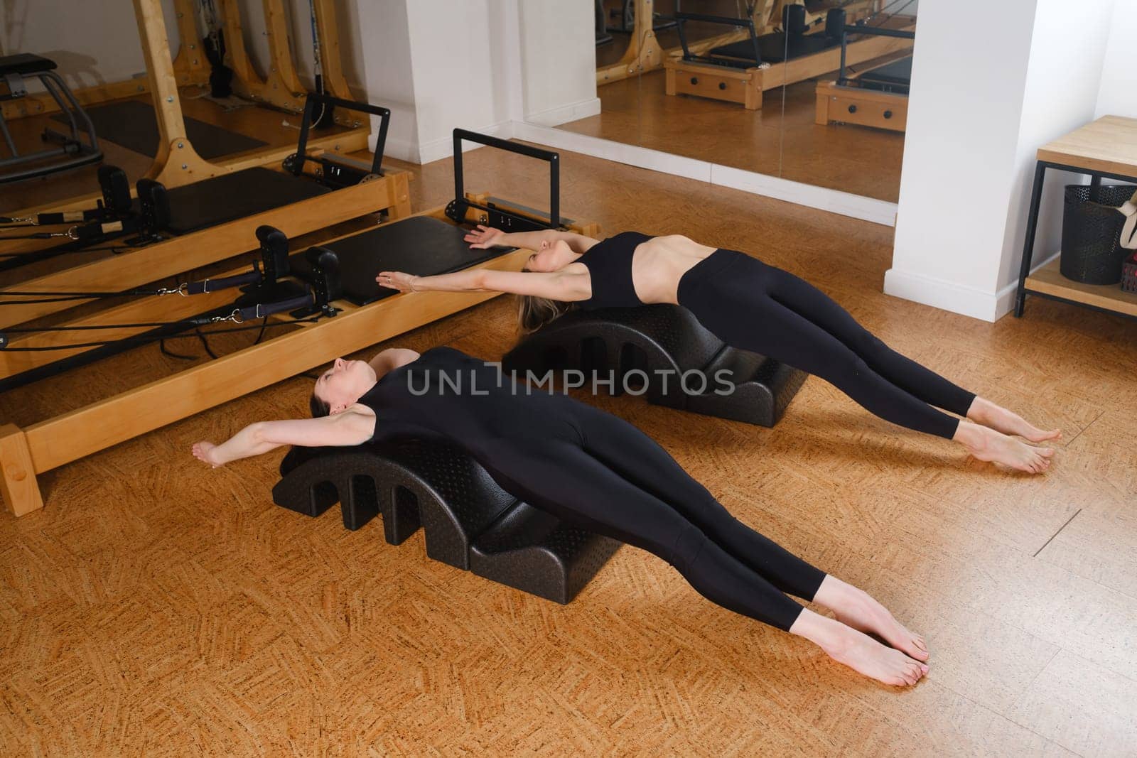 Two women are practicing physical fitness exercises on a Pilates machine in a room with hardwood flooring. Their movements showcase a beautiful balance of art and exercise