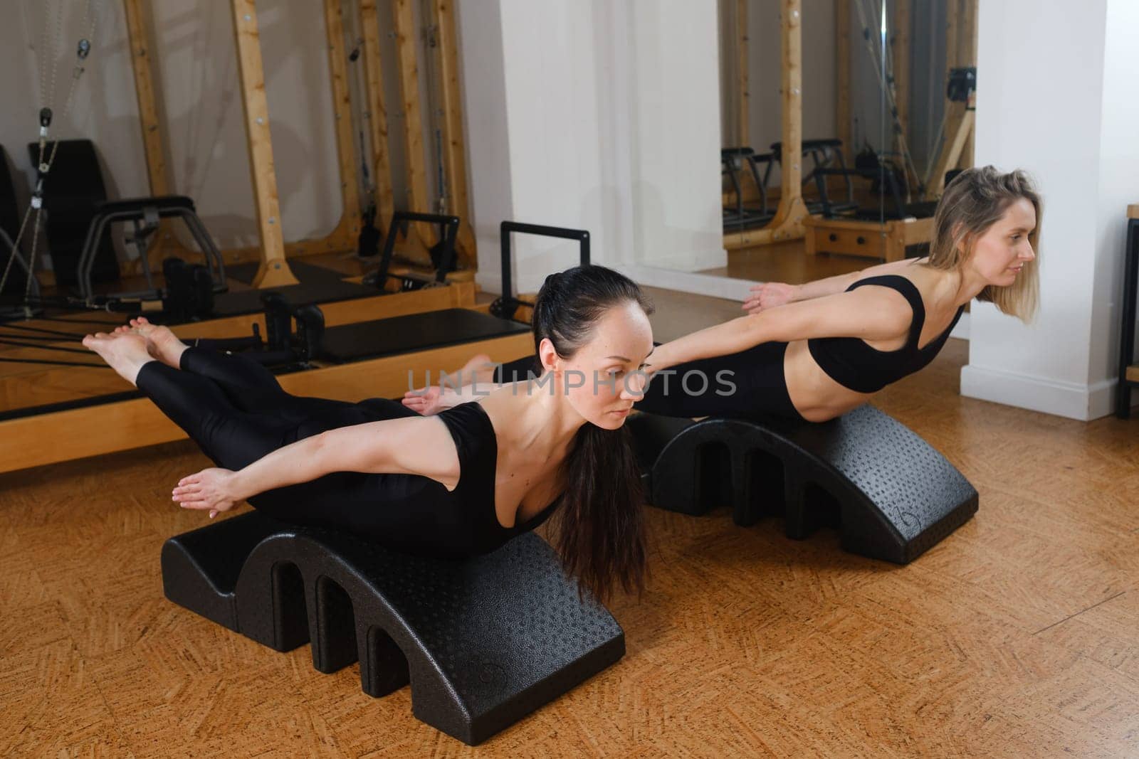 Two women are practicing physical fitness exercises on a Pilates machine in a room with hardwood flooring. Their movements showcase a beautiful balance of art and exercise