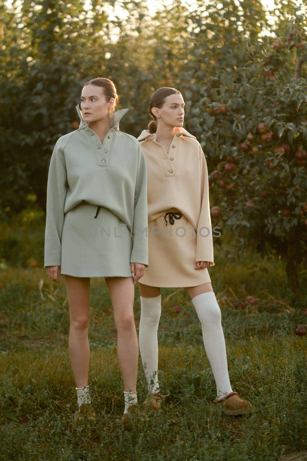 Two models in stylish tracksuits pose in apple orchard at sunset . The model is wearing a light-colored tracksuit and skirt.