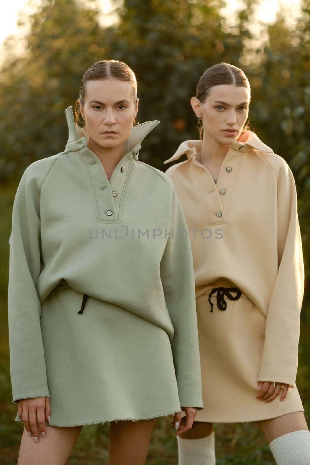 Two models in stylish tracksuits pose in apple orchard at sunset . The model is wearing a light-colored tracksuit and skirt.